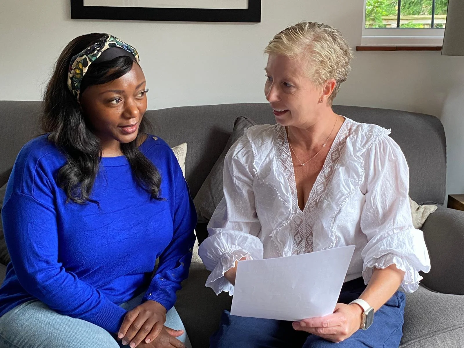 Two women sitting on a couch engaged in conversation, one holding a CV and talking through how best to structure her new CV.