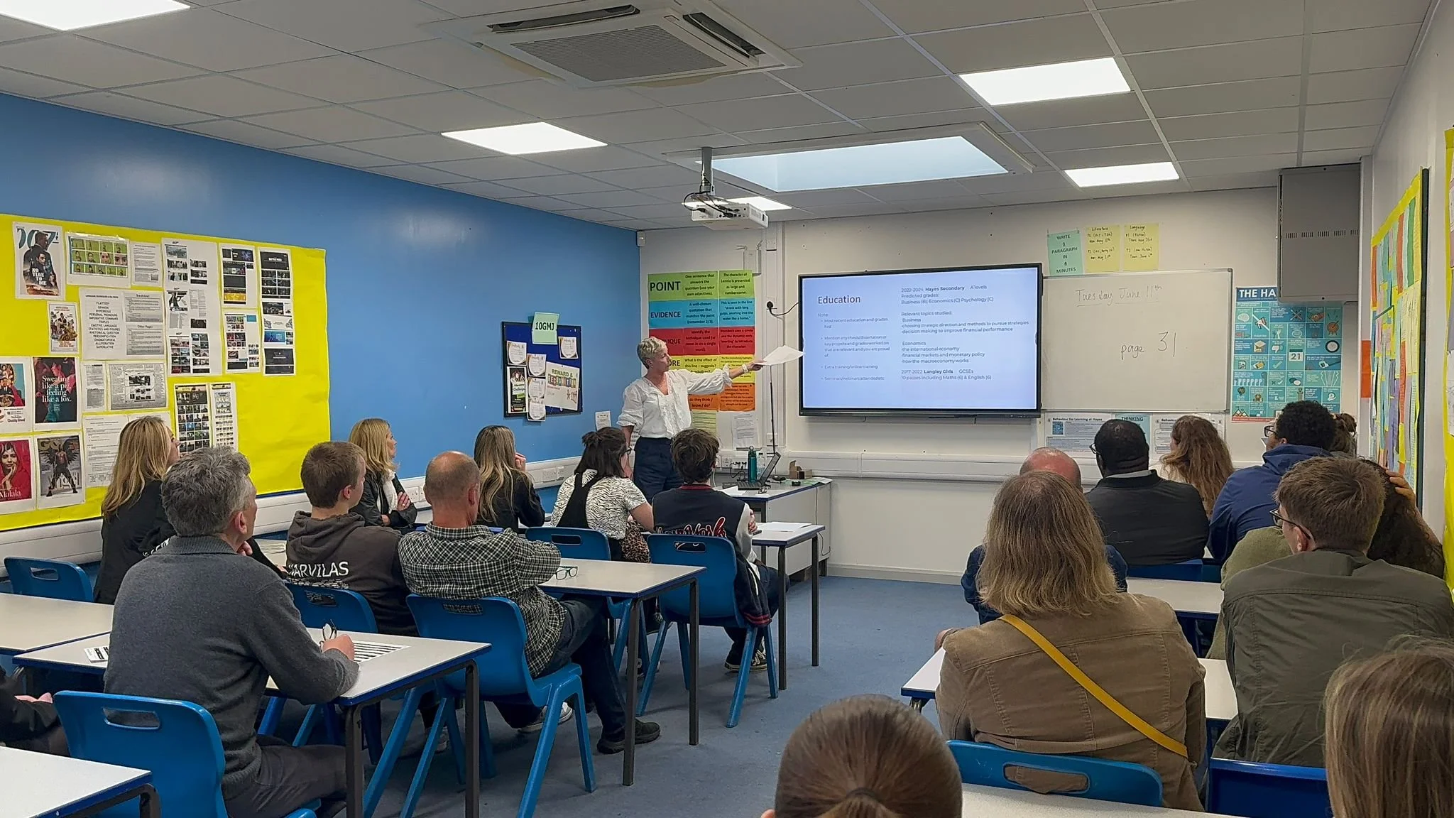 Classroom with students sitting at desks facing a teacher presenting slides on a large screen, with educational posters on the walls.