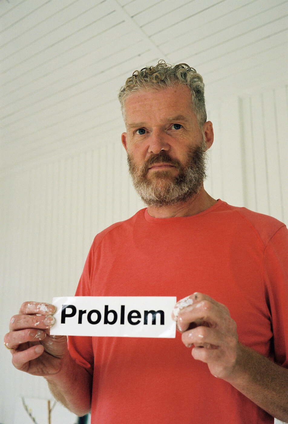 John Kørner in red shirt holding a sign that says 'Problem' with a serious expression.