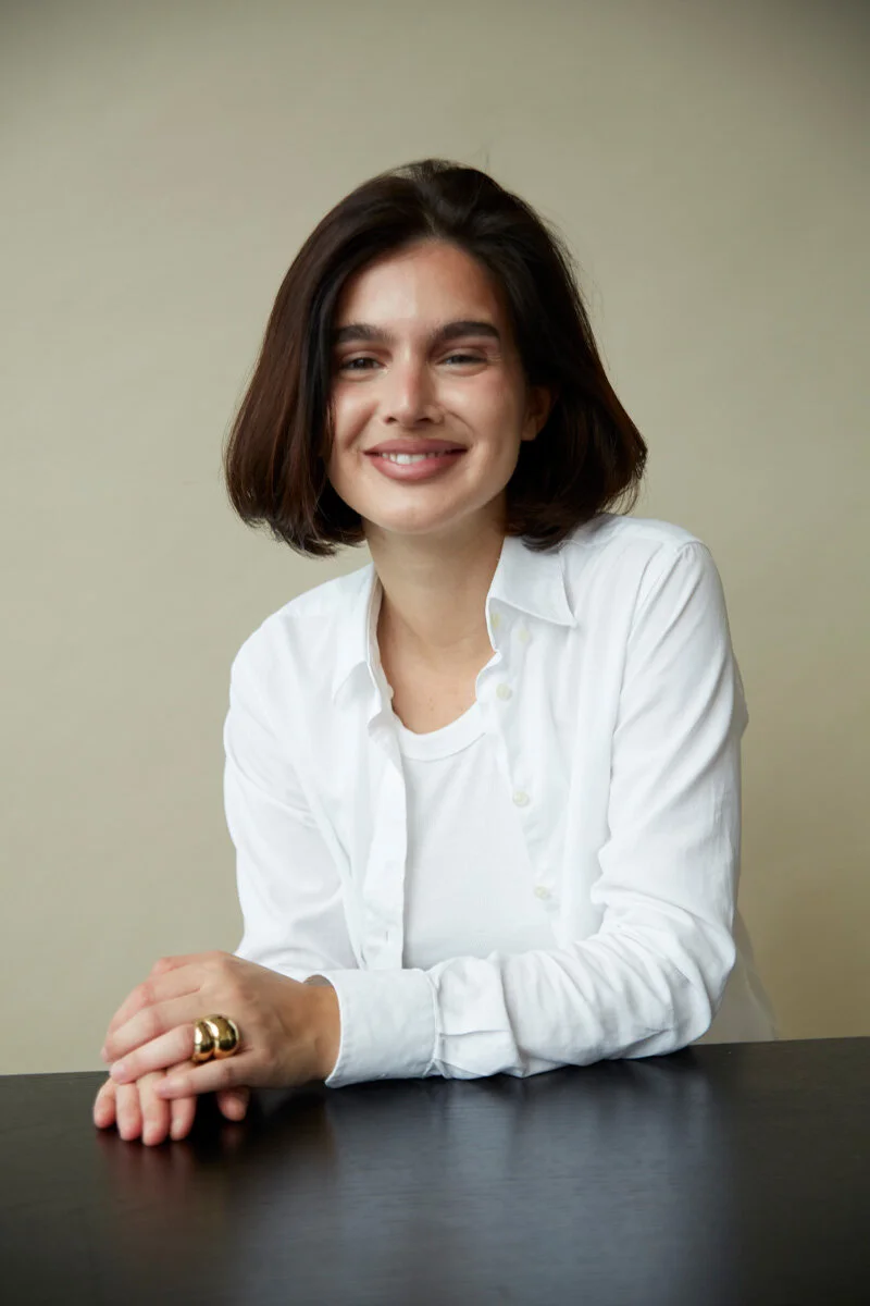 Smiling person with short brown hair wearing a white shirt, seated at a table.