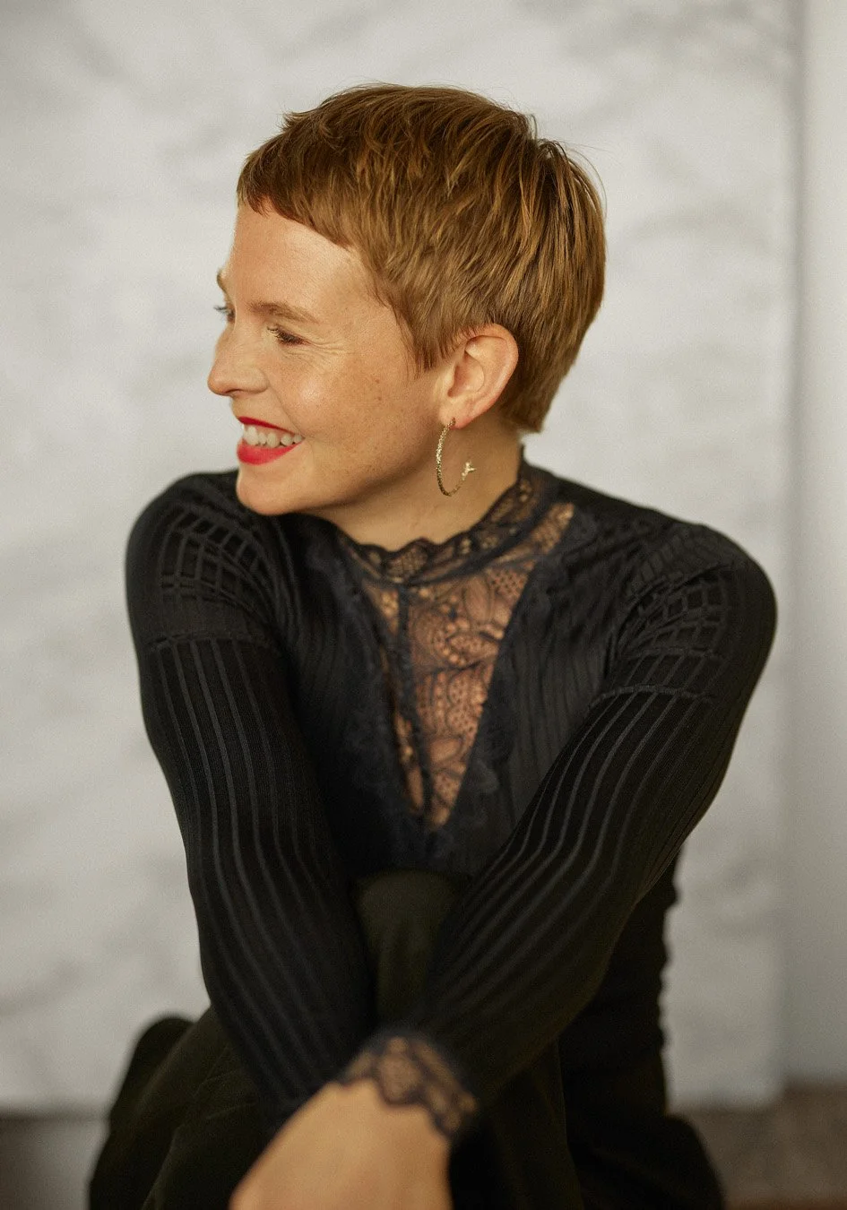 Person smiling sideways, wearing a black lace top, short hair, and hoop earrings.
