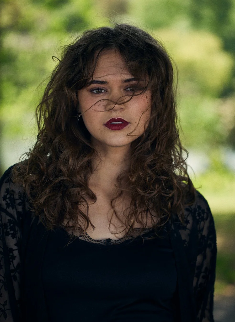 Portrait of a person with curly hair and wearing a black top, outdoors with a blurred natural background.
