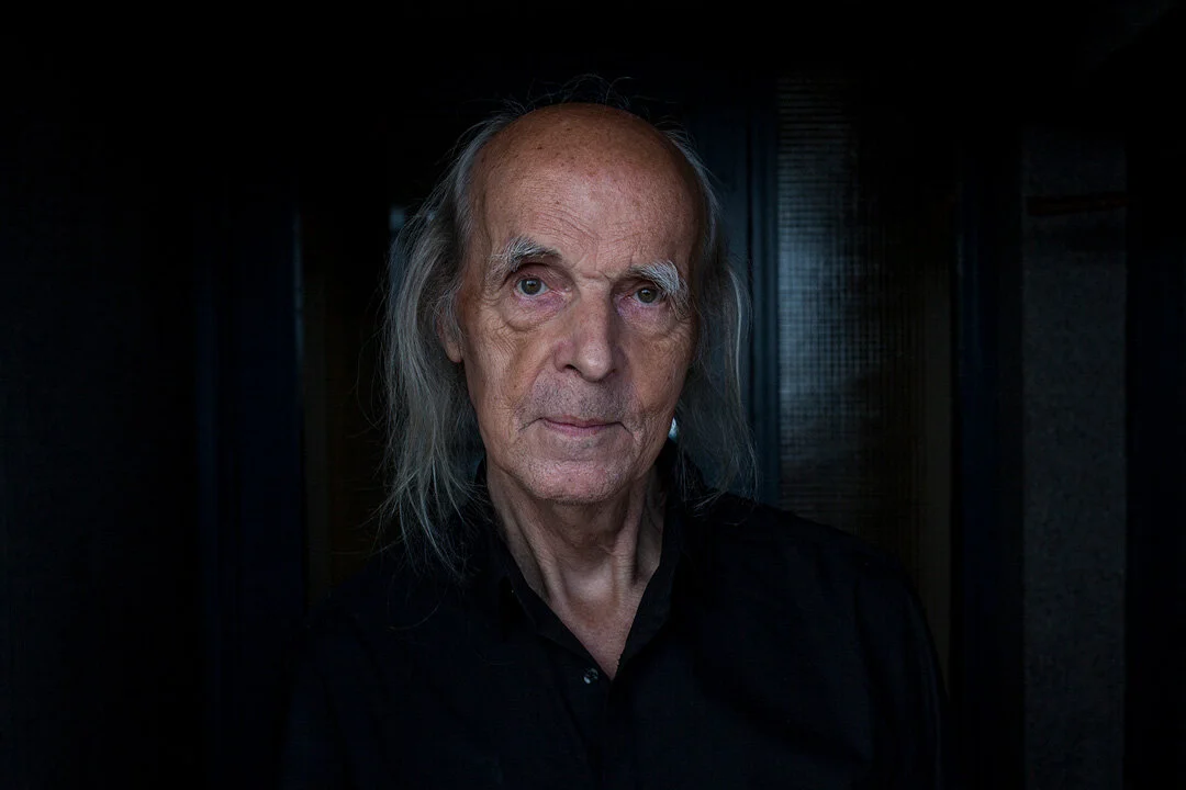 Elderly man with long hair and a black shirt against a dark background.