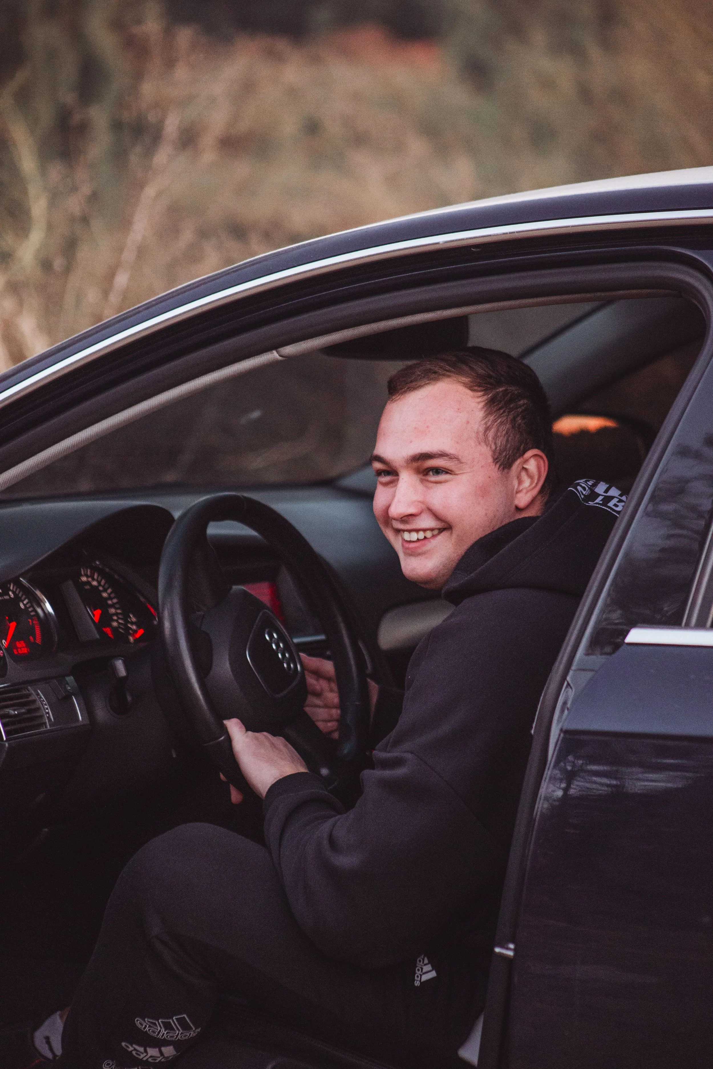 A smiling young man with short brown hair sitting in the driver's seat of a car, holding the steering wheel, with a view of the car's dashboard and a wooded landscape outside the window.