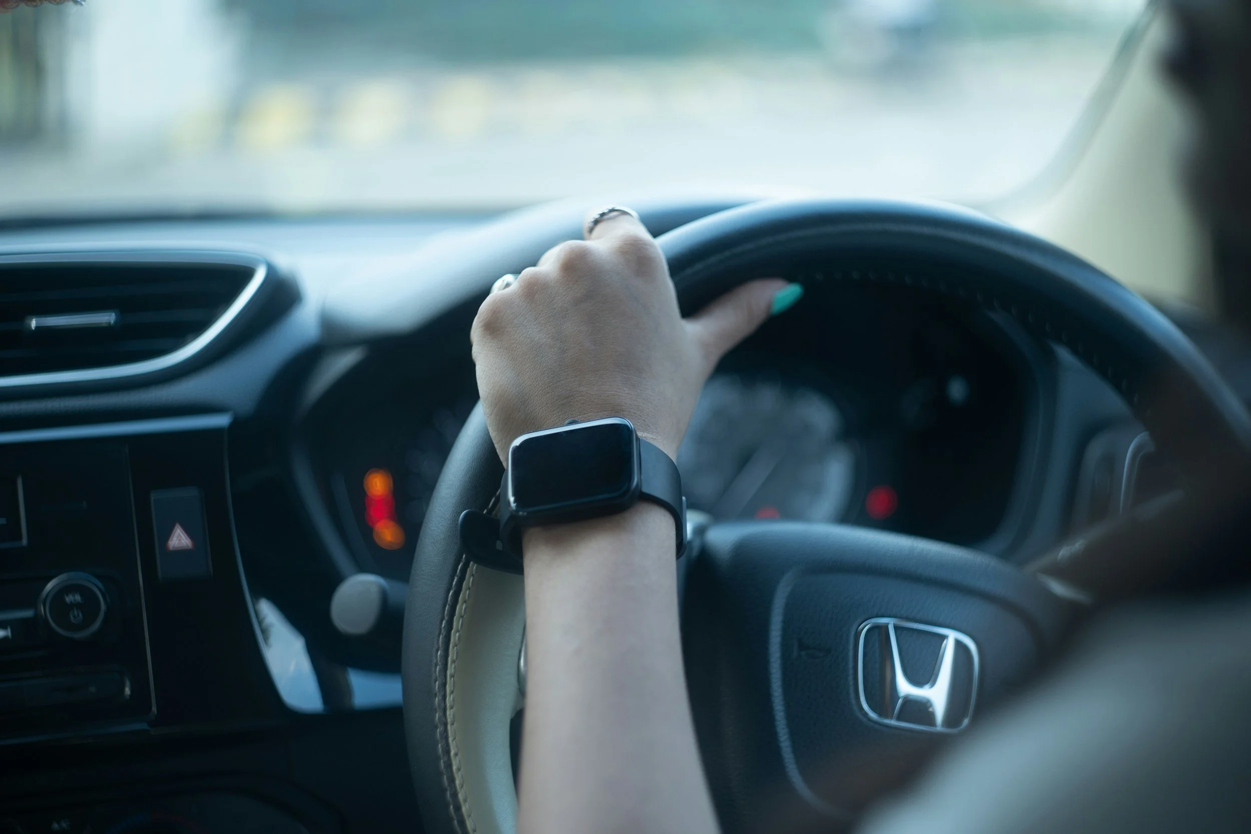A person driving a car, holding the steering wheel with their left hand, wearing a smartwatch and a ring, with visible interior dashboard and air vent.