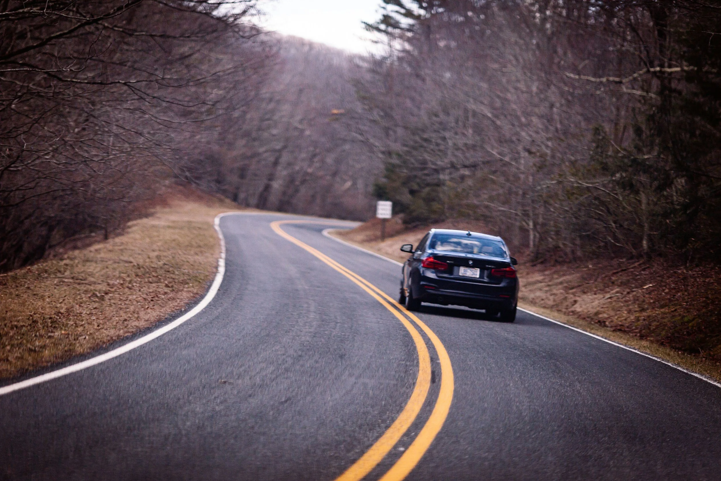 A black car driving on a winding road through a wooded area with leafless trees and a small sign on the side of the road.