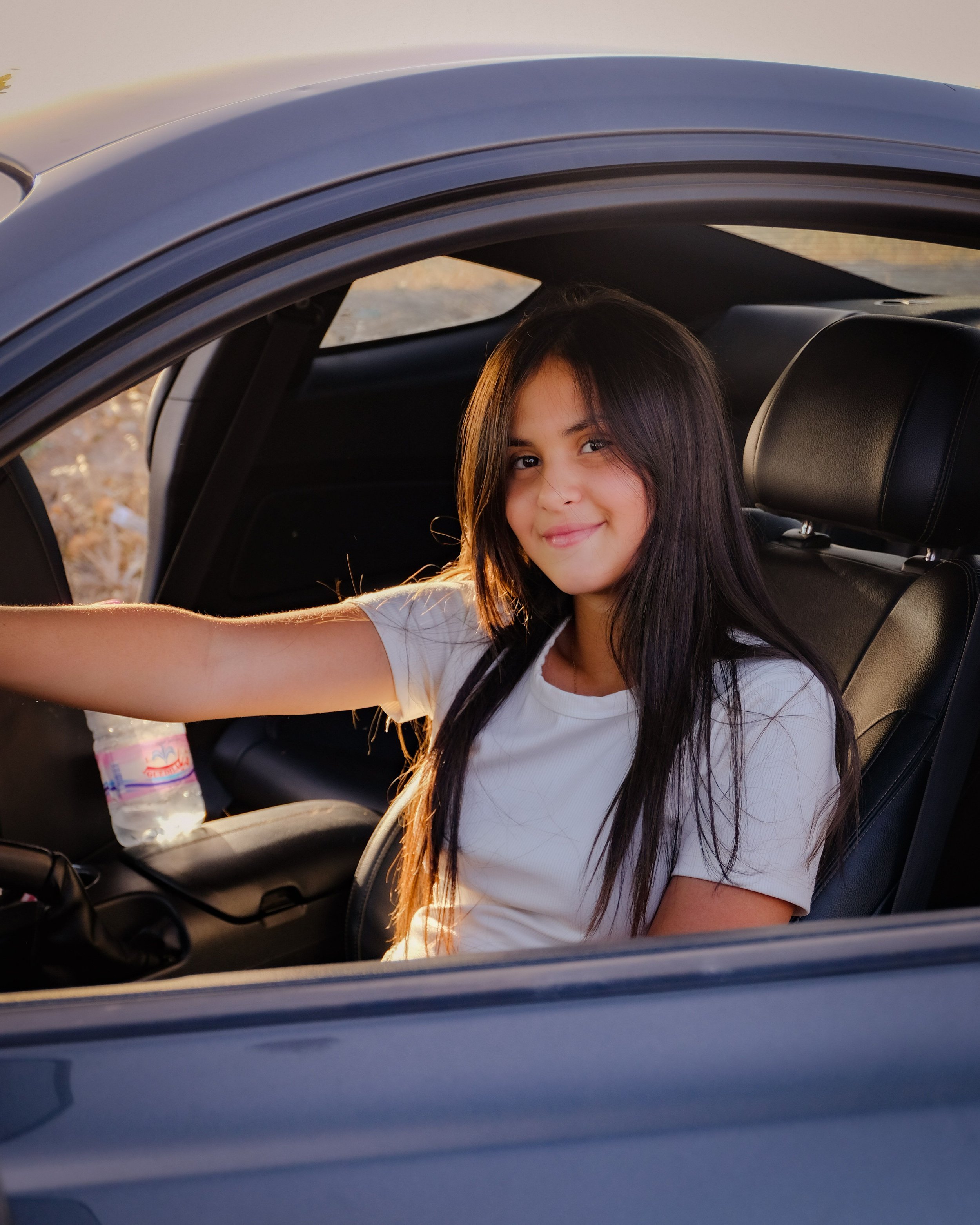 A young girl with long dark hair sitting in the back seat of a car, smiling at the camera. The car door is open, and a plastic water bottle is on the seat next to her. The background shows a sunset or sunrise outside the car.