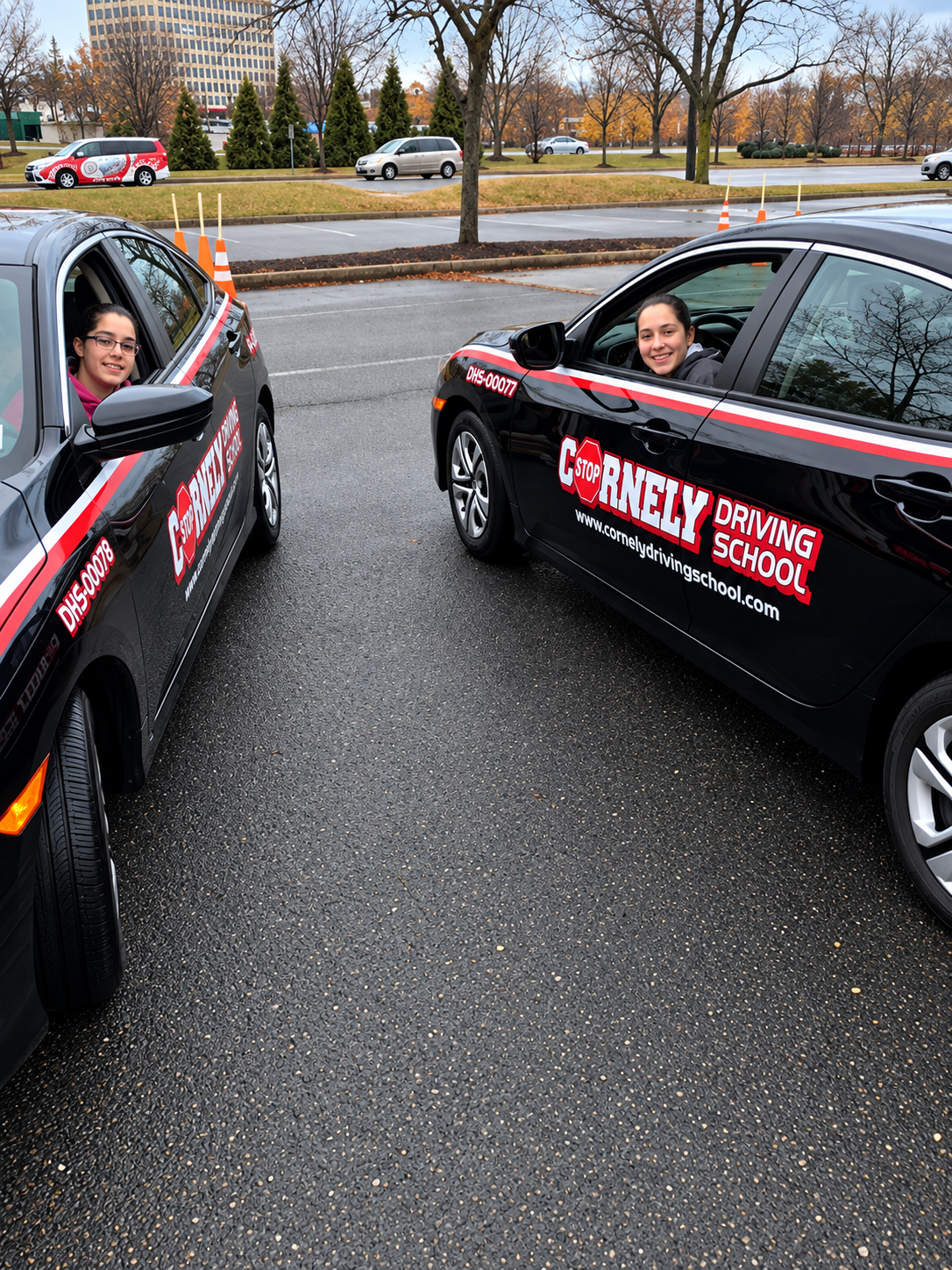 Two young women in driver training cars from Cornely Driving School, parked facing each other in a wet parking lot on a cloudy day, with trees and buildings in the background.