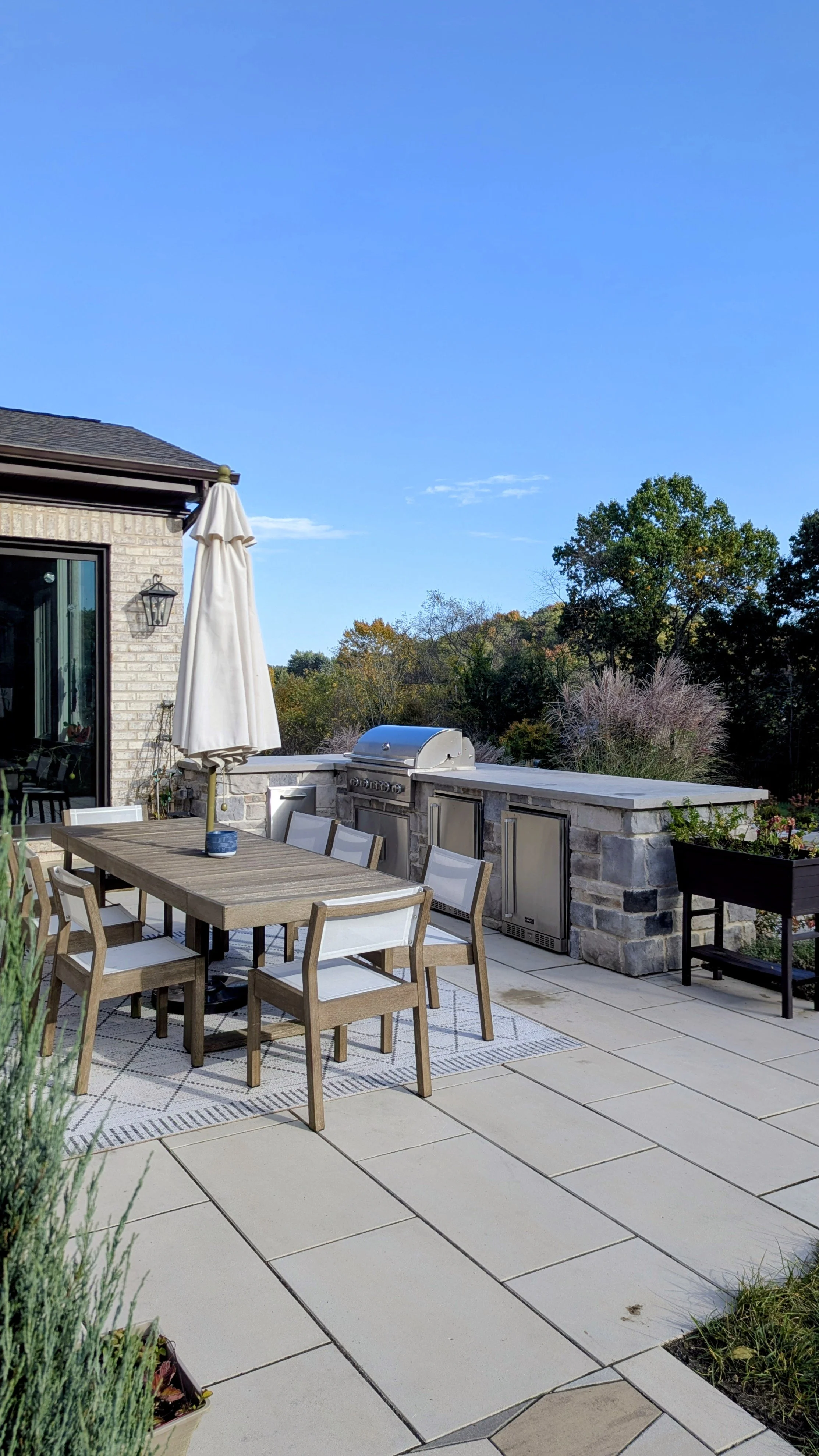 Outdoor patio with a wooden dining table, chairs, an umbrella, a built-in stainless steel grill, and panoramic trees in the background under a clear blue sky.