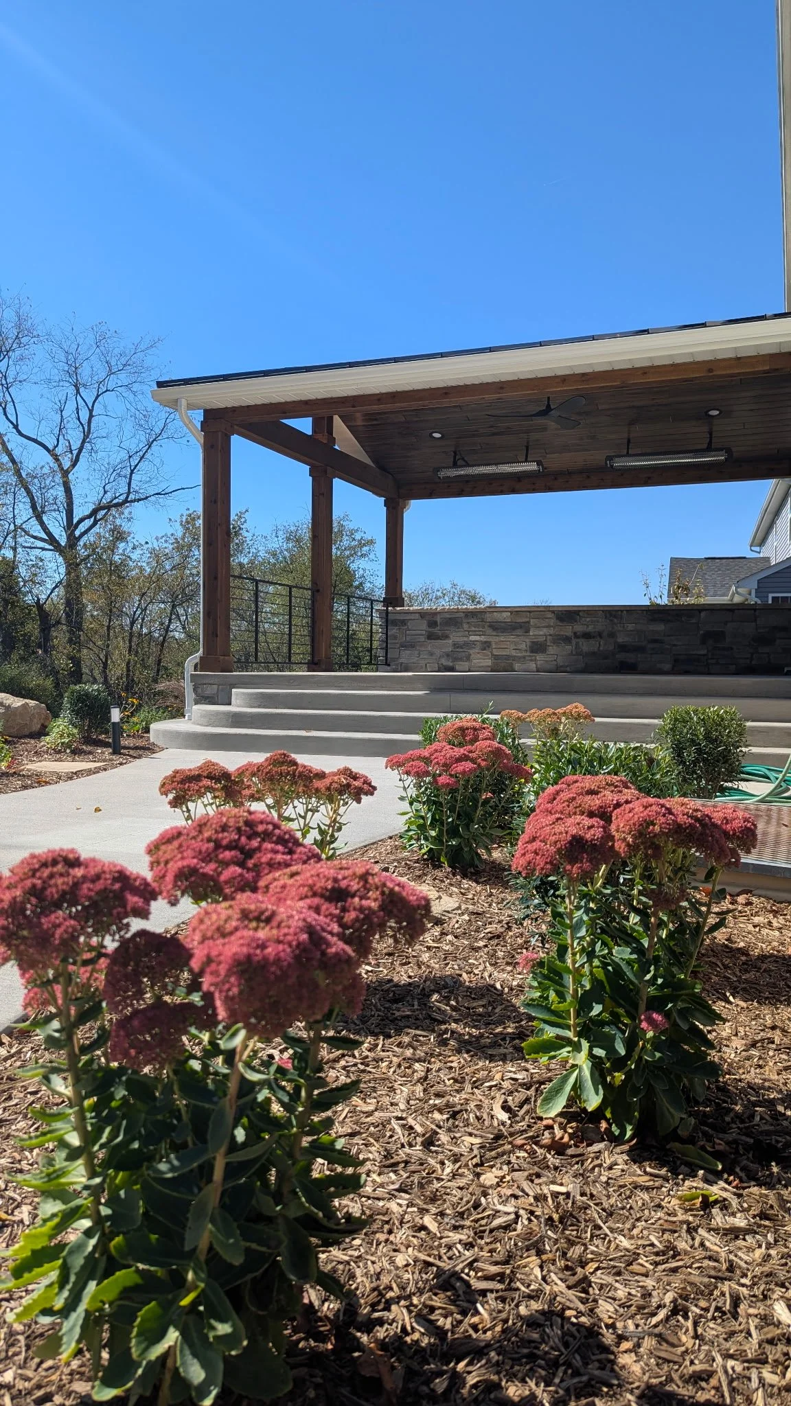A backyard patio with concrete steps leading up to a covered area supported by wooden posts, pink flowering plants in the foreground, a stone wall behind the steps, and a clear blue sky with a faint rainbow in the background.