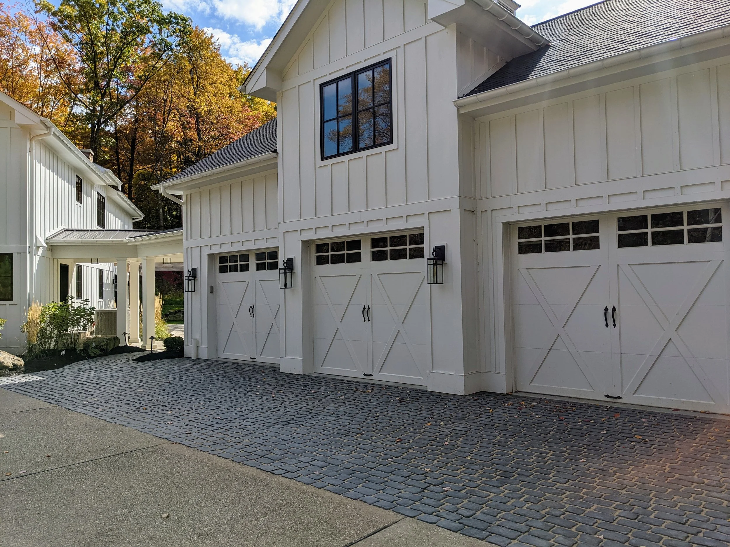 White house with two garages, black framed windows, and a cobblestone driveway set against a backdrop of trees with fall foliage under a partly cloudy sky.