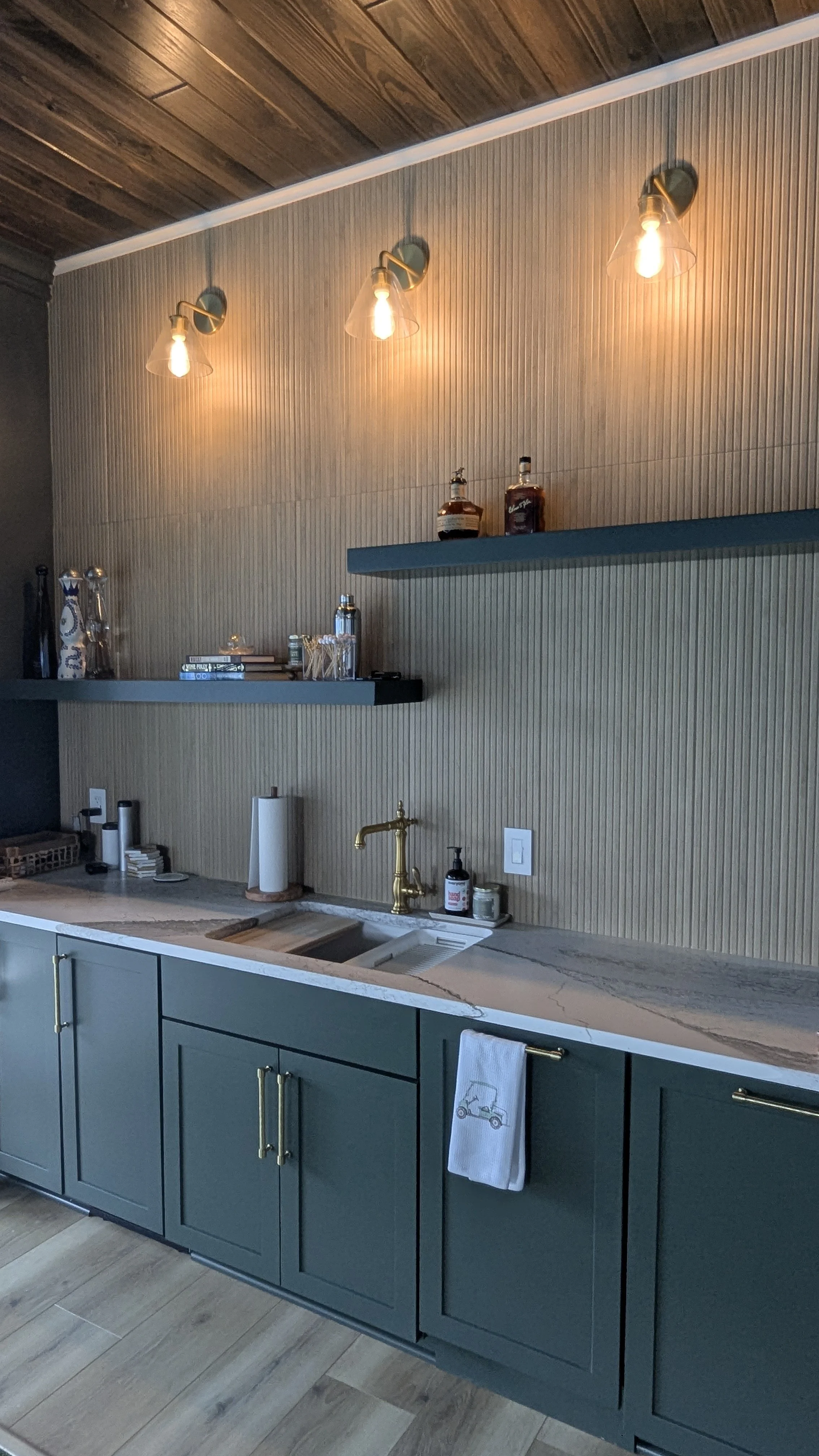 Modern kitchen with green cabinets, marble countertop, gold faucet, beige textured wall, wooden ceiling, and three wall-mounted lights with glass shades.