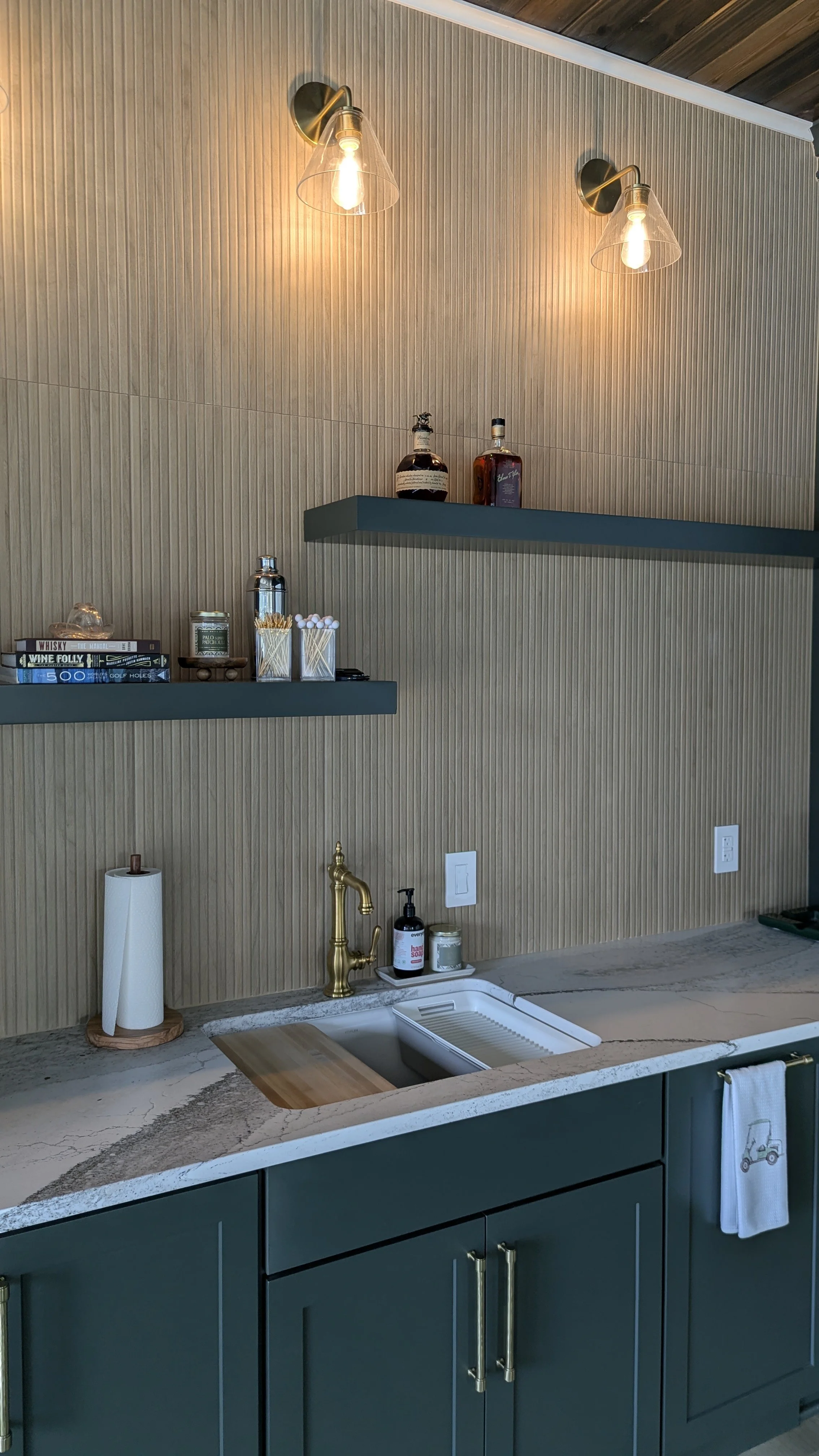 Kitchen countertop with a sink, gold faucet, and various items including a paper towel roll, soap, and decorative jars, with two floating shelves holding liquor bottles and books, and wall-mounted lights above.