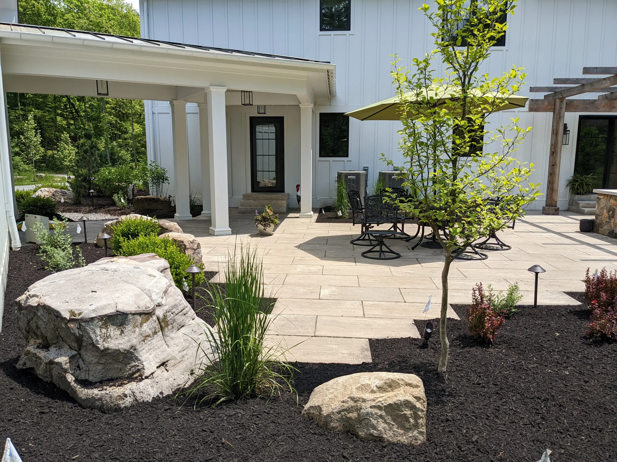 A modern house exterior featuring a small patio area with black metal furniture, a green umbrella, landscaped garden with rocks and plants, and a white building with black-framed windows and doors.