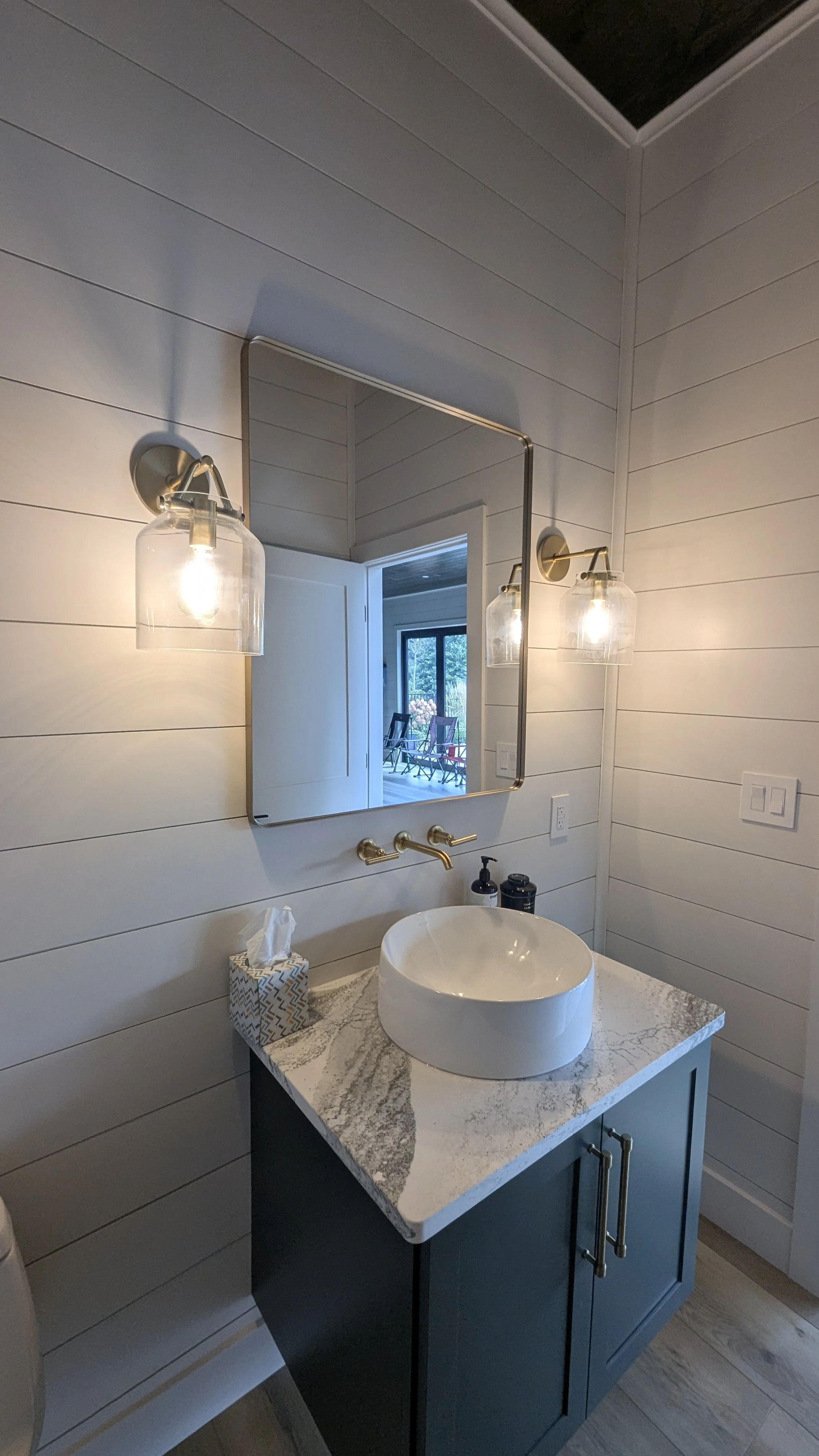 Modern bathroom with white shiplap walls, a gold-framed mirror, and two wall-mounted glass light fixtures. There is a vessel sink on a marble countertop with a blue cabinet underneath. A tissue box and soap dispenser are on the counter. A window with
