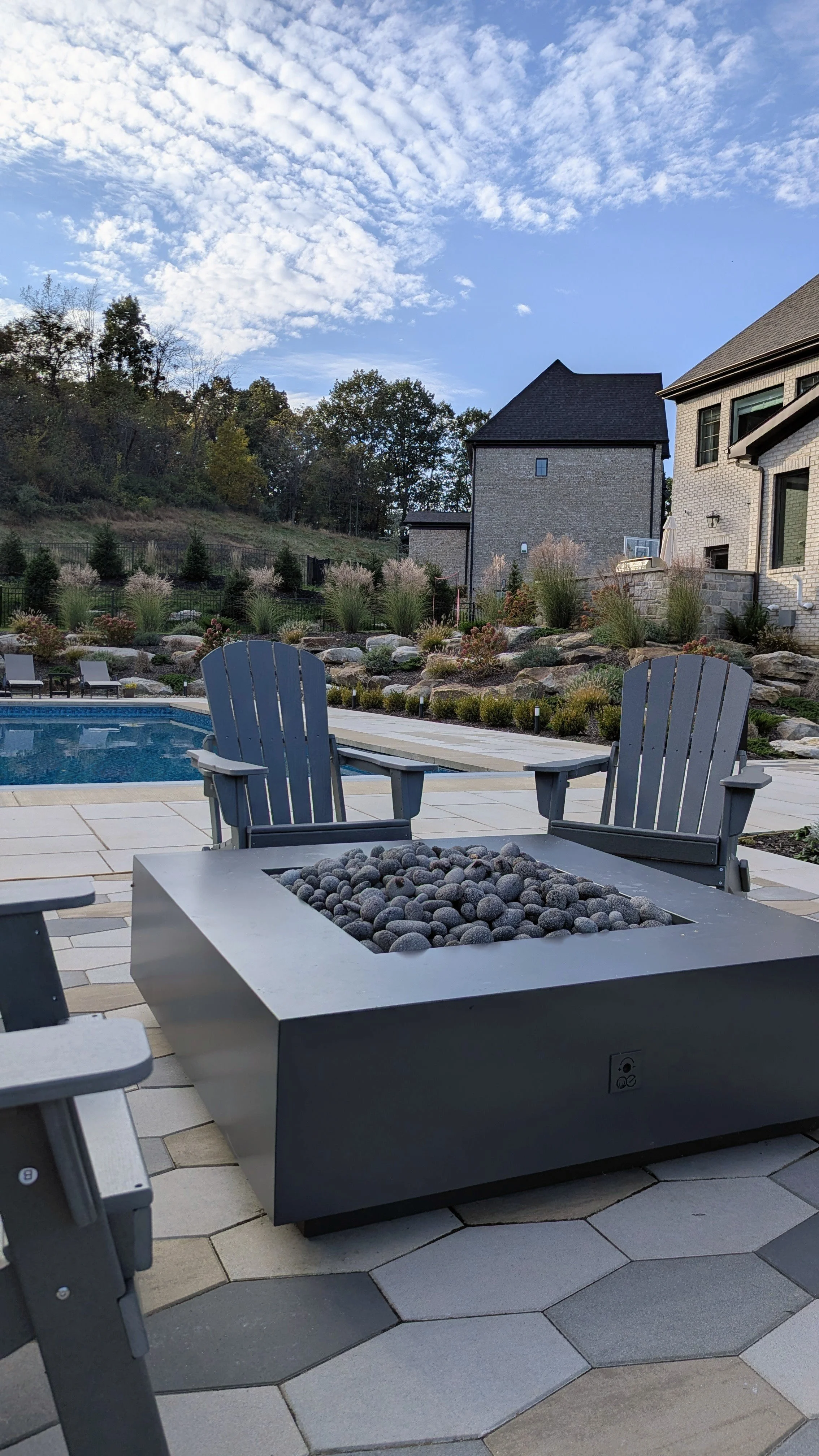 A backyard patio area with a fire pit filled with decorative rocks, seating, a swimming pool, and landscaped garden with bushes and rocks. In the background, there are two houses and a hill covered with trees. The sky is mostly cloudy with patches of
