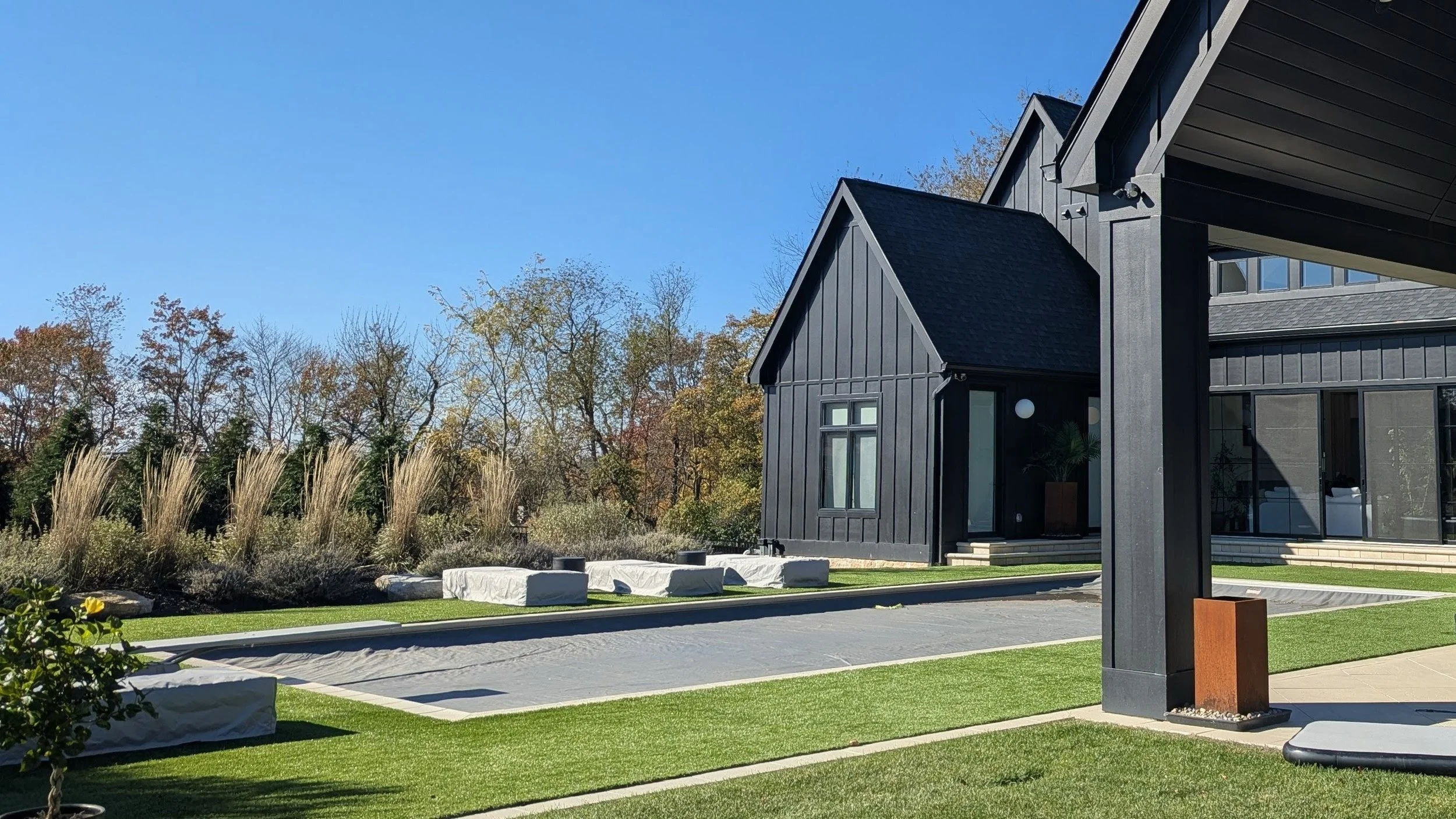 Modern black house with sharp triangular roofs, large windows, and outside seating area, set in a landscaped yard with green grass, plants, and trees under a clear blue sky.