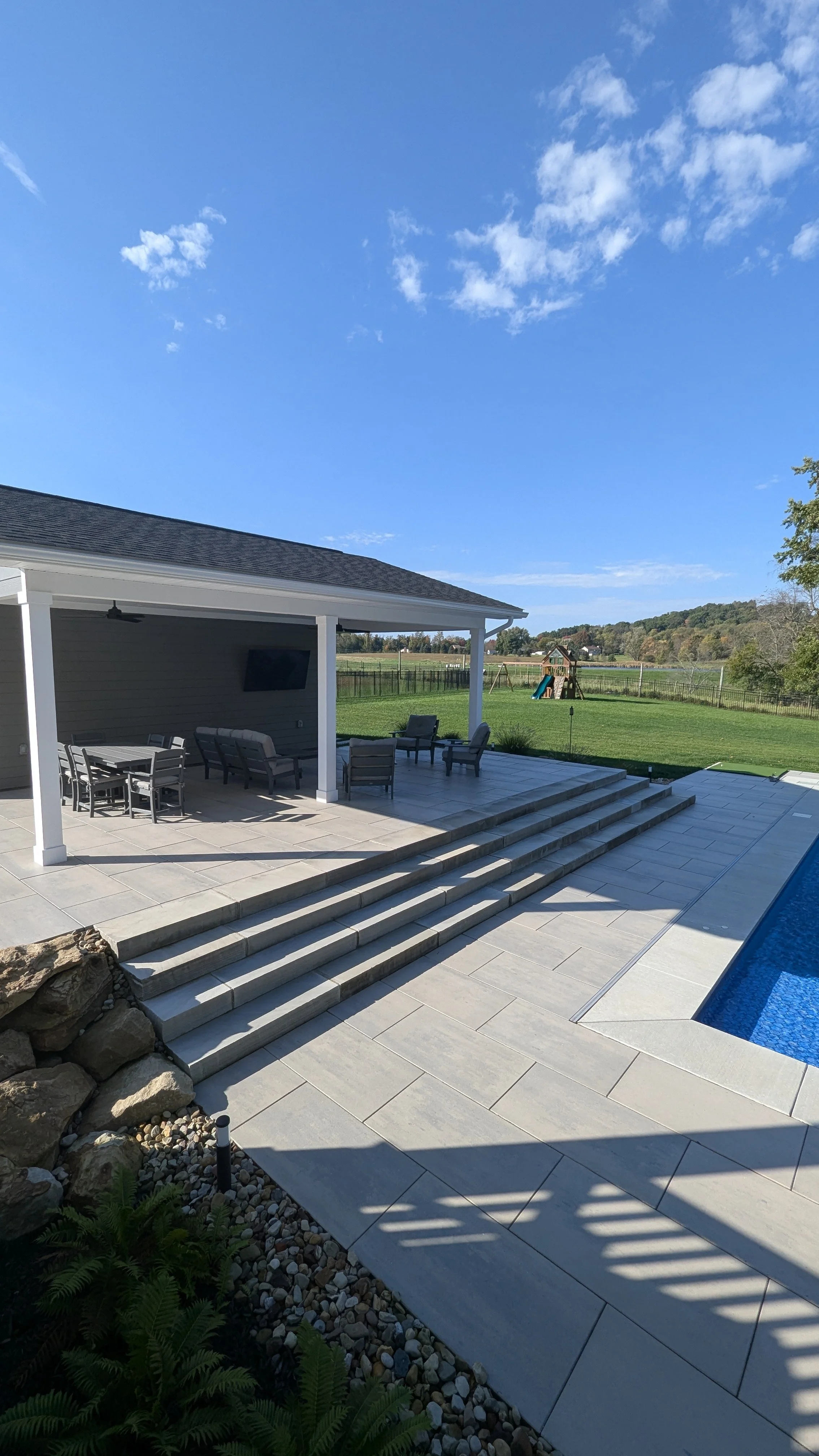 Backyard patio with outdoor furniture, steps leading down to a grassy yard, playset in the distance, and a clear blue sky with clouds.