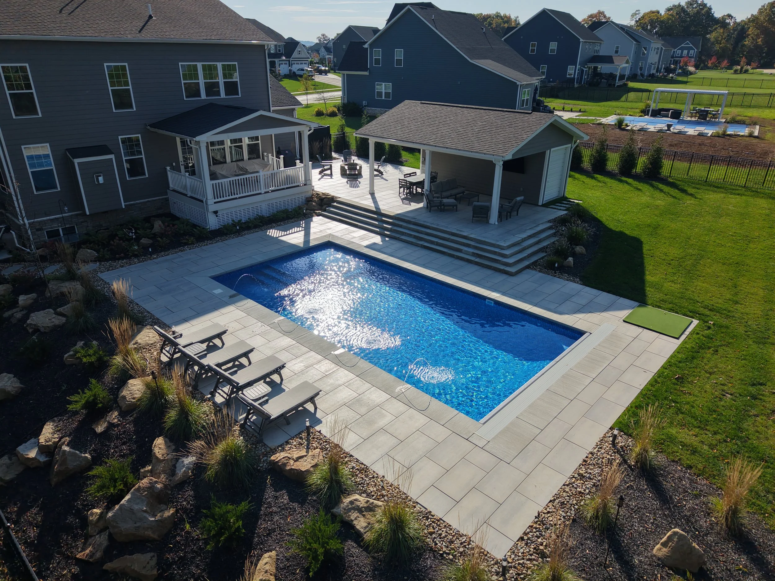 A backyard with a swimming pool, outdoor seating area, a covered patio, and a nearby swimming pool with loungers and a pergola, surrounded by a fenced yard and neighboring houses.