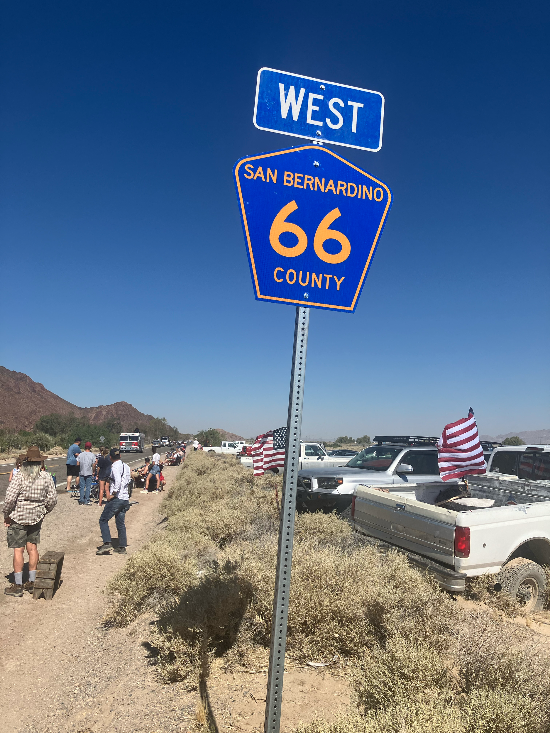 A sign indicating West on U.S. Route 66 in San Bernardino County, with people and parked cars on a desert roadside under a clear blue sky.