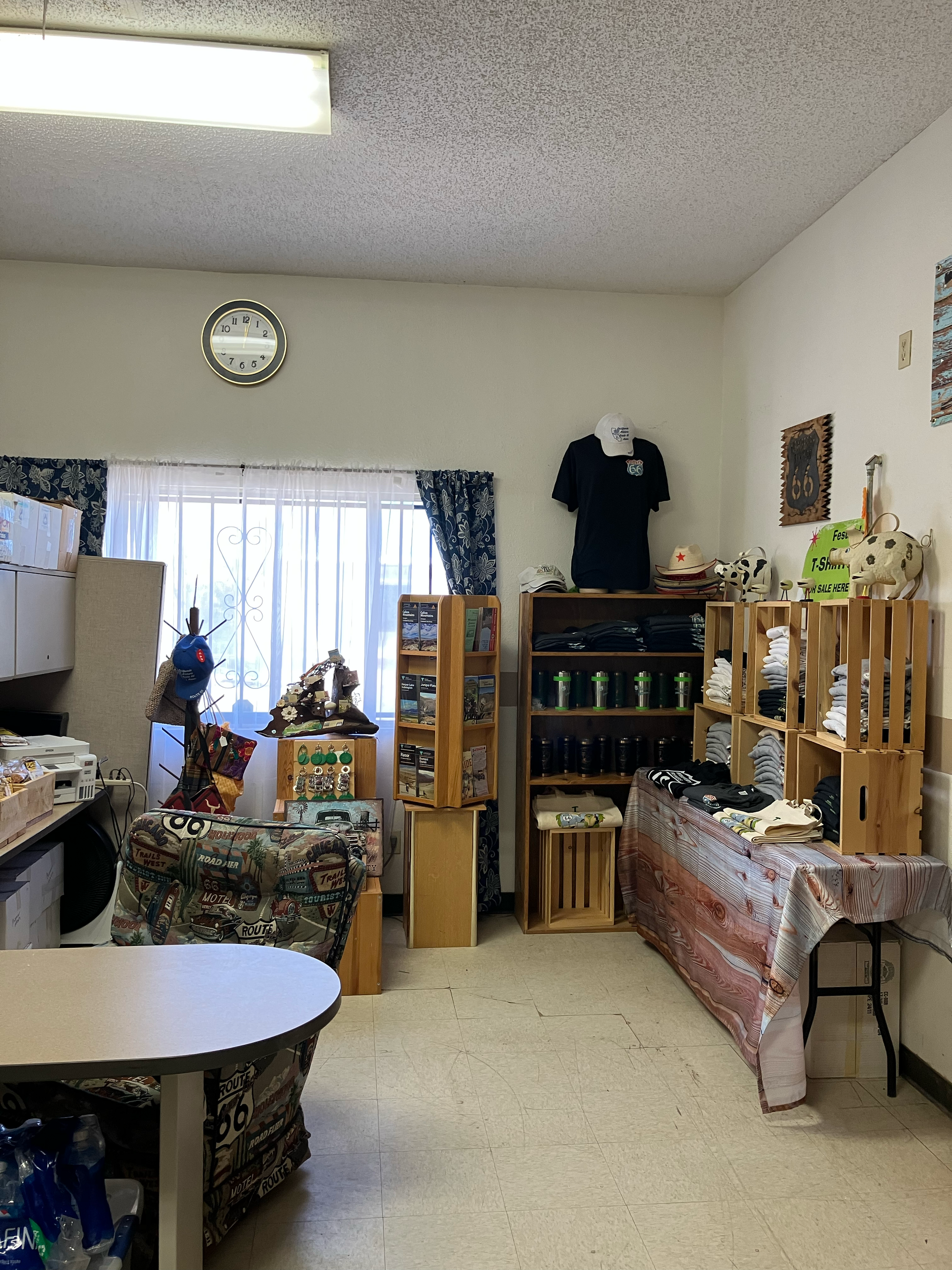 Room with a wall-mounted clock, a window with curtains, shelves with hats and sunglasses, a display of glass bottles, and tables covered with cloths displaying various items.