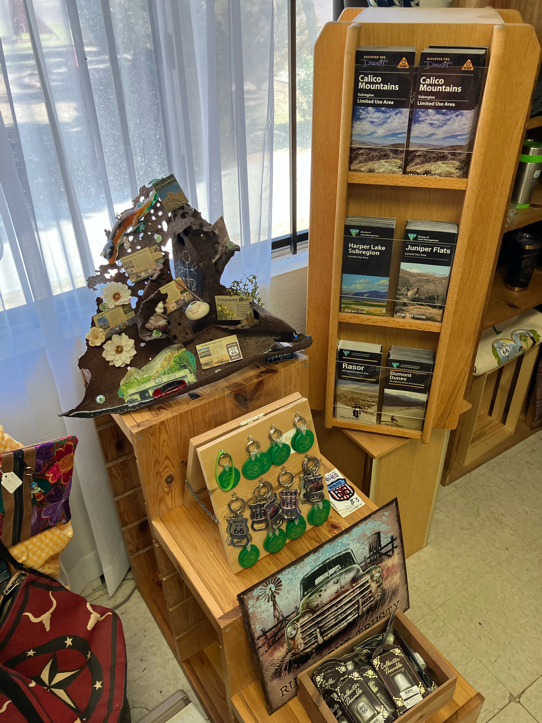 Display of souvenirs and brochures with a wooden rack of brochures about mountains and landscape destinations, a table with keychains and truck-themed items, and a decor piece shaped like a mountain with small decorative items, with a sheer white cur