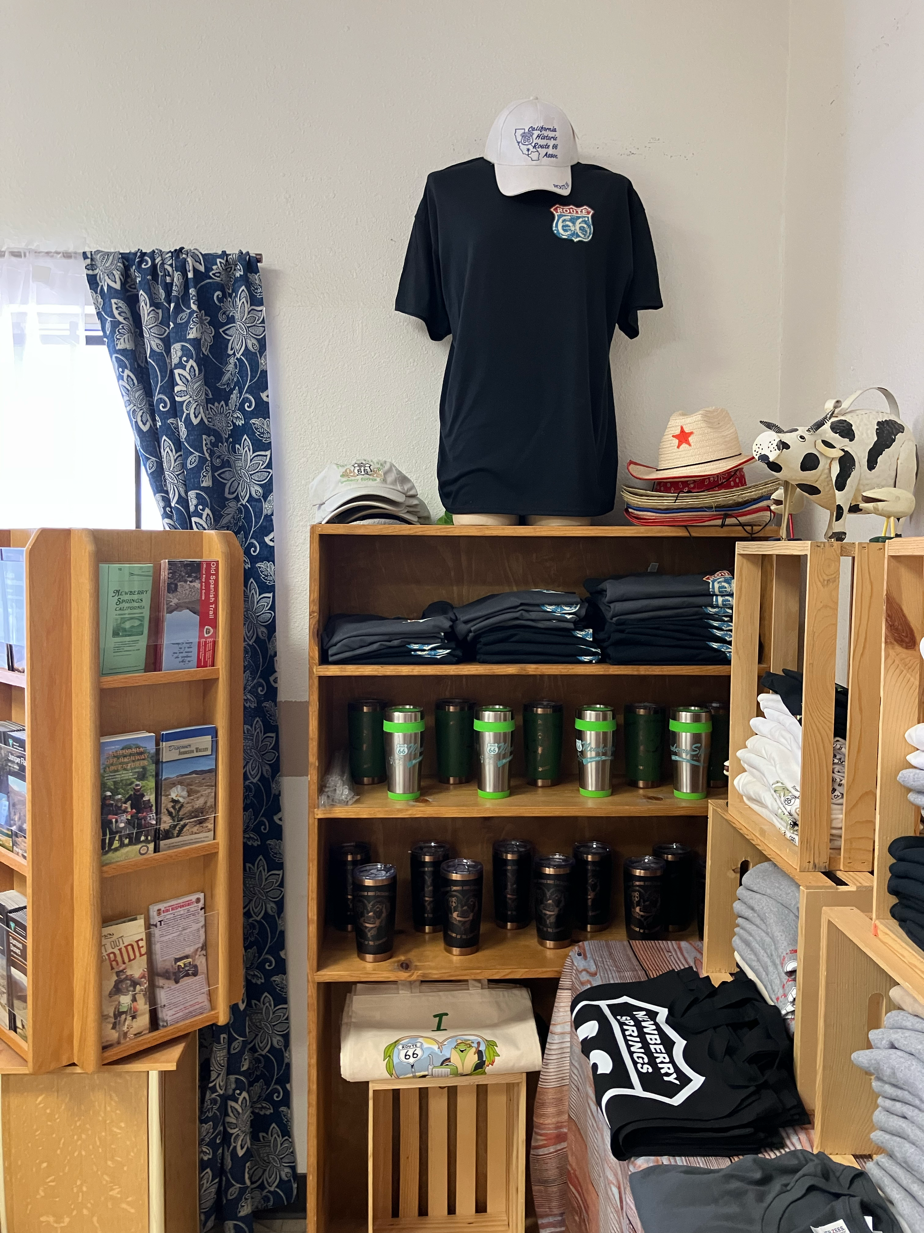 Display shelf with tiered wooden compartments holding green and black drink tumblers, folded shirts, and miscellaneous items including hats and a plush cow toy in a retail setting.