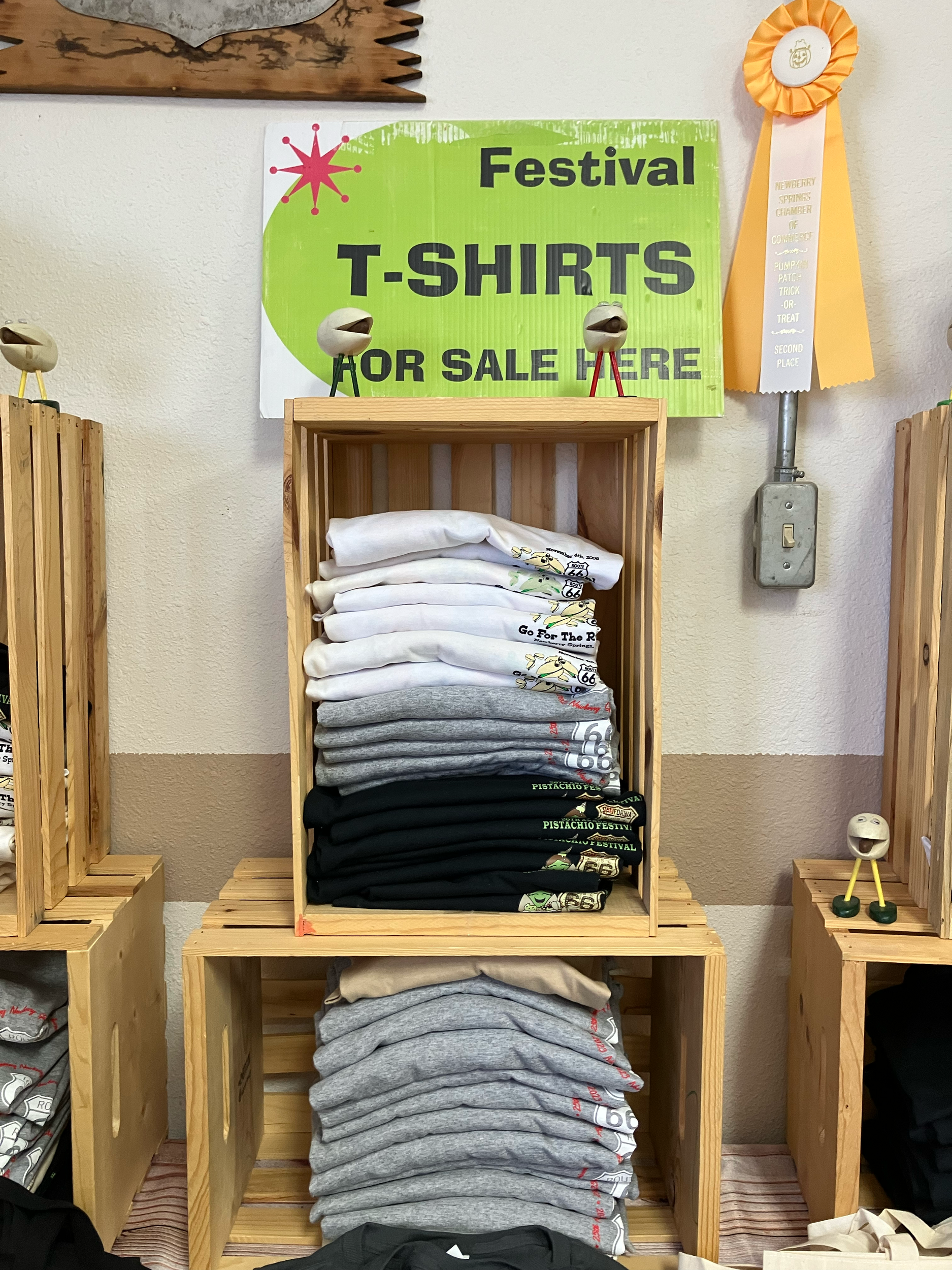 Display of t-shirts and sweatshirts for sale at a fundraising event, with a green sign reading 'Fundraiser T-Shirts for Sale Here,' and stacked clothing arranged on wooden shelves.