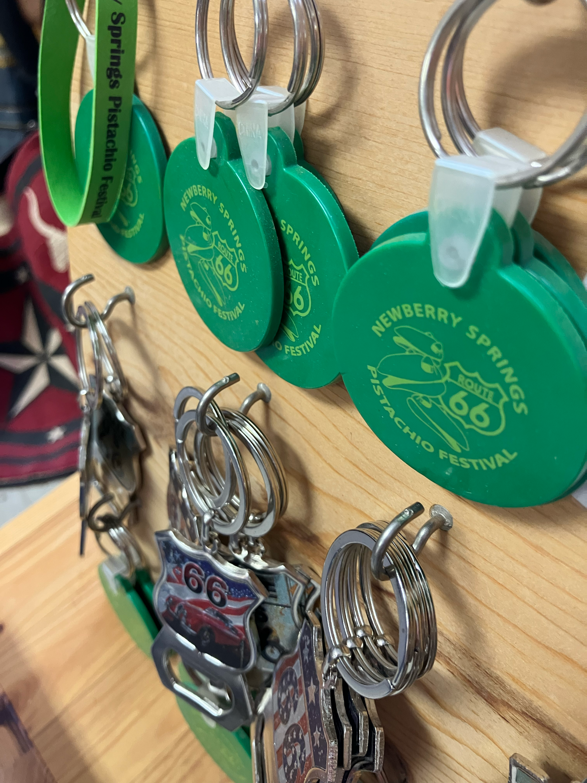 Multiple keychains and pins on a wooden surface, including round green keychains with a pistachio nut graphic and the text 'Nebraska Springs Pistachio Festival,' and metal pins with the Route 66 emblem.