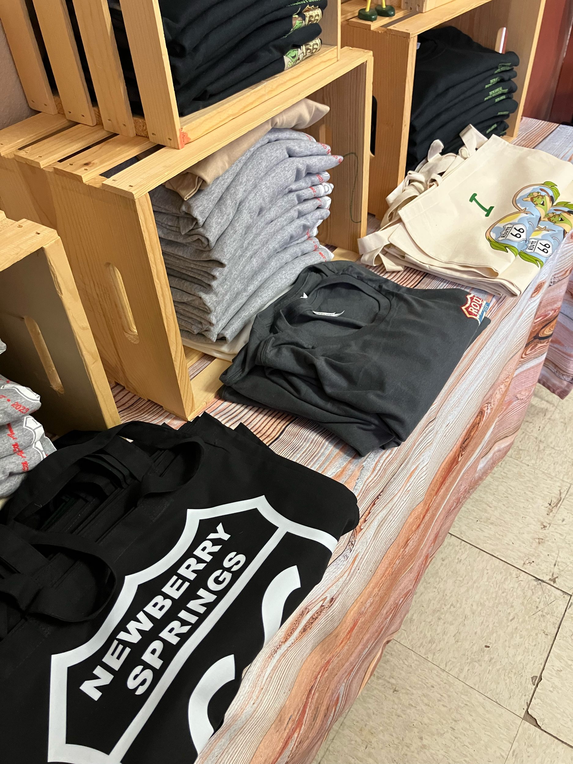 Various clothing items including black, gray, and beige folded hoodies or sweatshirts, and a black bag with a white shield logo and text reading "Newberry Spruings" on a wooden table.