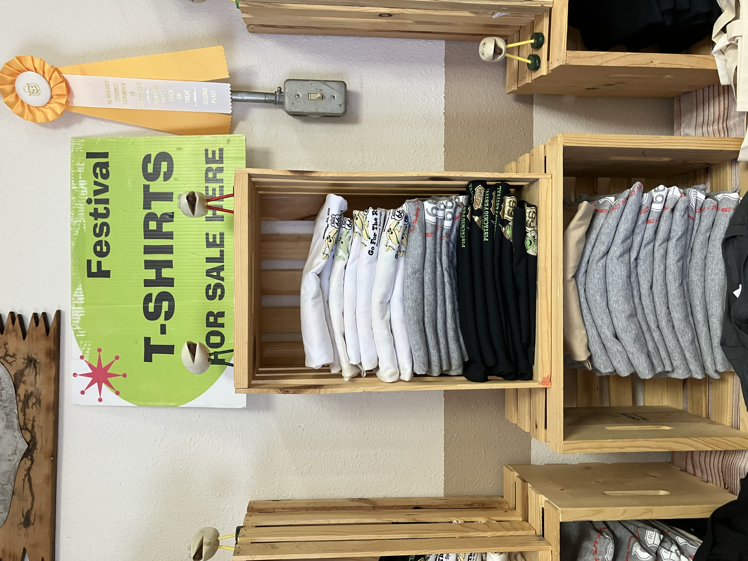Display of t-shirts and sweatshirts for sale at a fundraising event, with a green sign reading 'Fundraiser T-Shirts for Sale Here,' and stacked clothing arranged on wooden shelves.