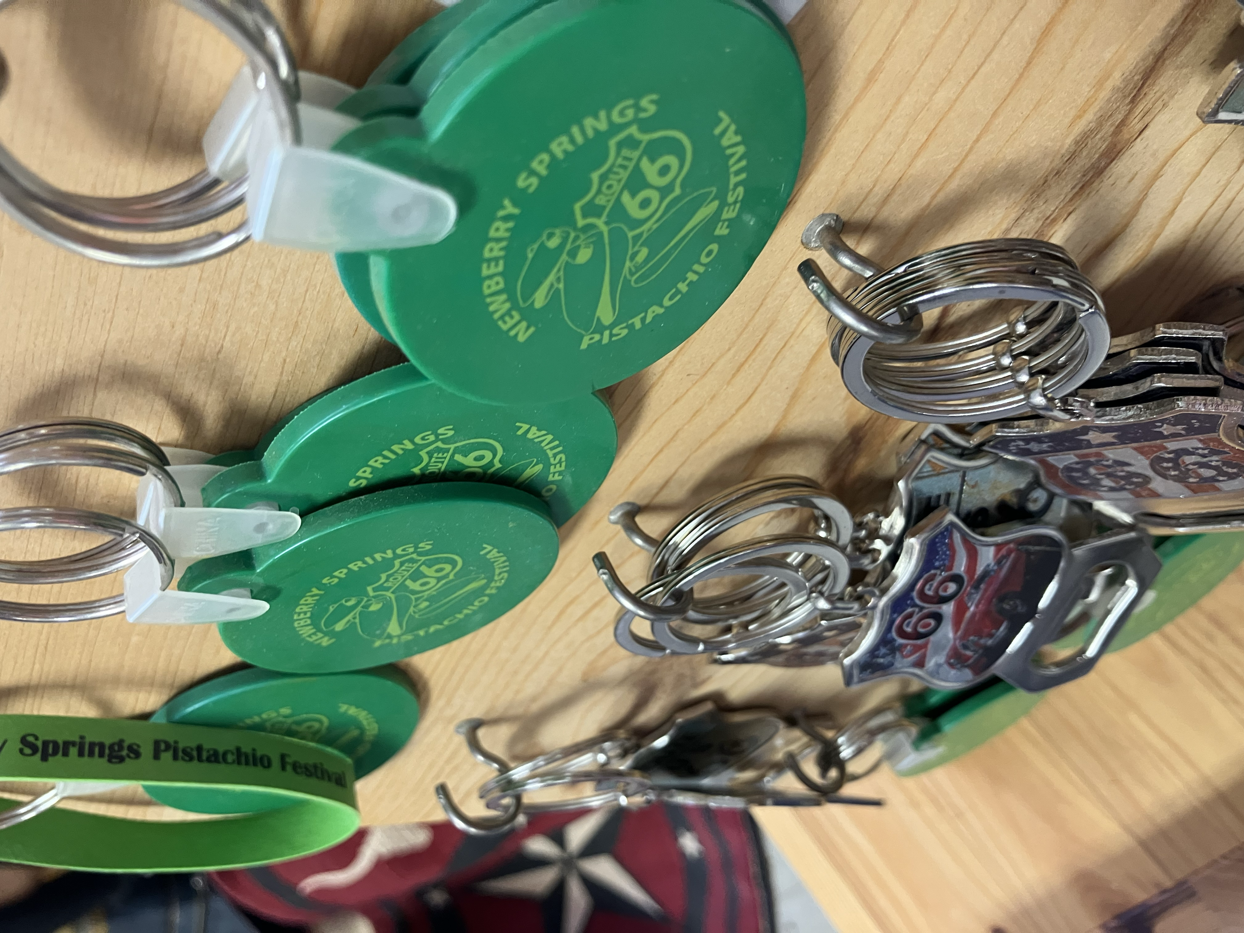 Multiple keychains and pins on a wooden surface, including round green keychains with a pistachio nut graphic and the text 'Nebraska Springs Pistachio Festival,' and metal pins with the Route 66 emblem.