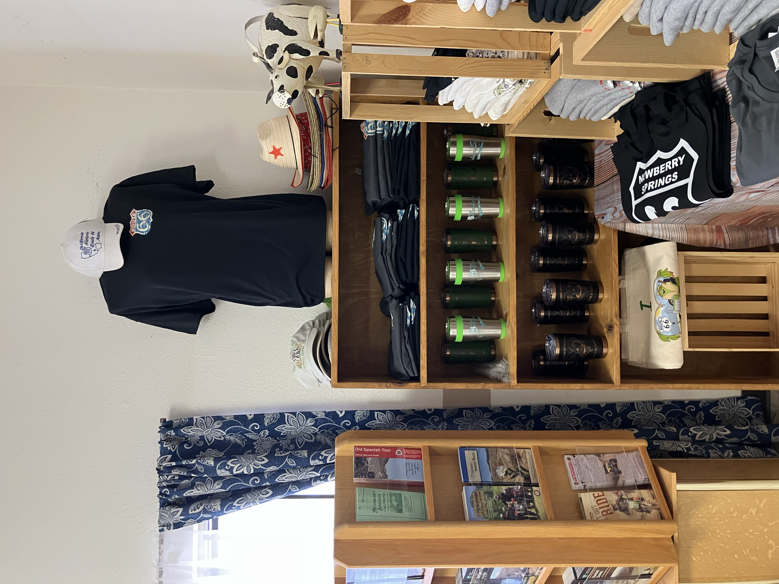 Display shelf with tiered wooden compartments holding green and black drink tumblers, folded shirts, and miscellaneous items including hats and a plush cow toy in a retail setting.