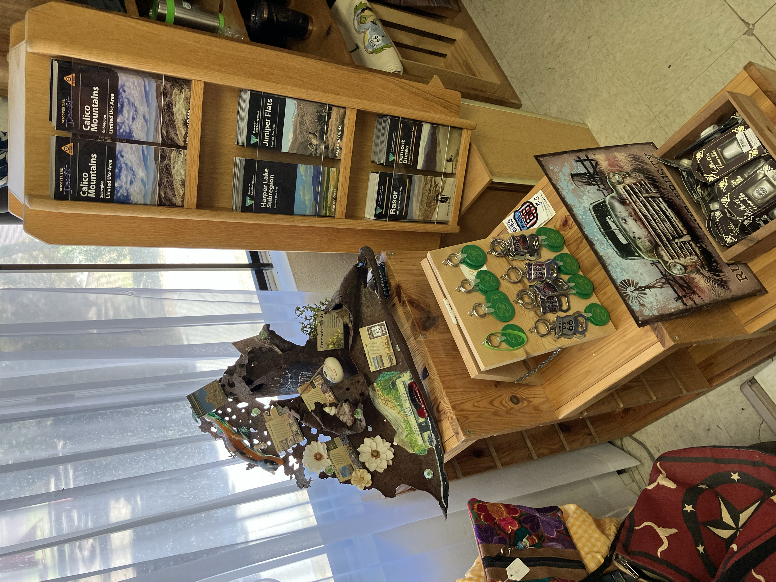 Display of souvenirs and brochures with a wooden rack of brochures about mountains and landscape destinations, a table with keychains and truck-themed items, and a decor piece shaped like a mountain with small decorative items, with a sheer white cur