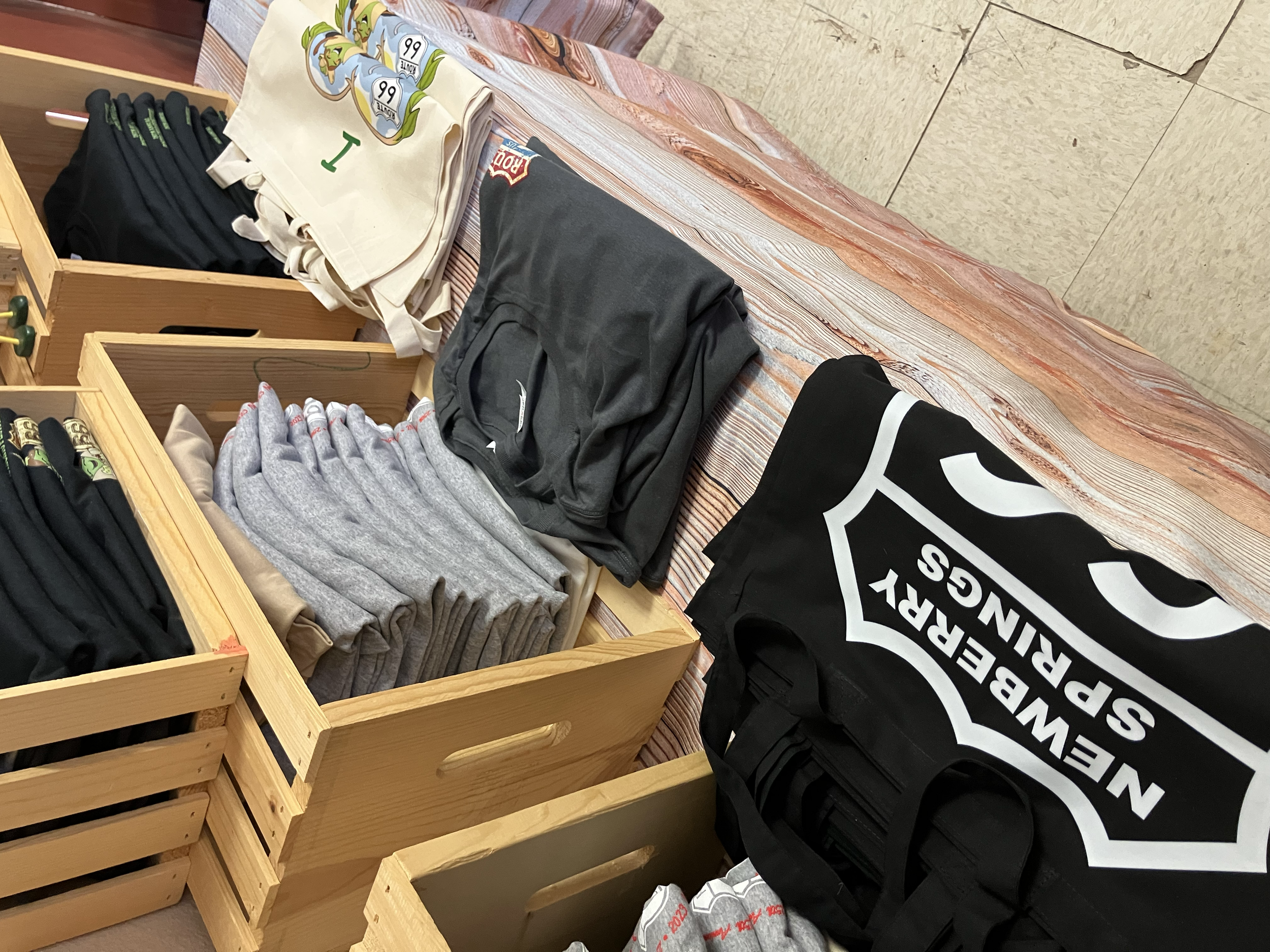 Various clothing items including black, gray, and beige folded hoodies or sweatshirts, and a black bag with a white shield logo and text reading "Newberry Spruings" on a wooden table.