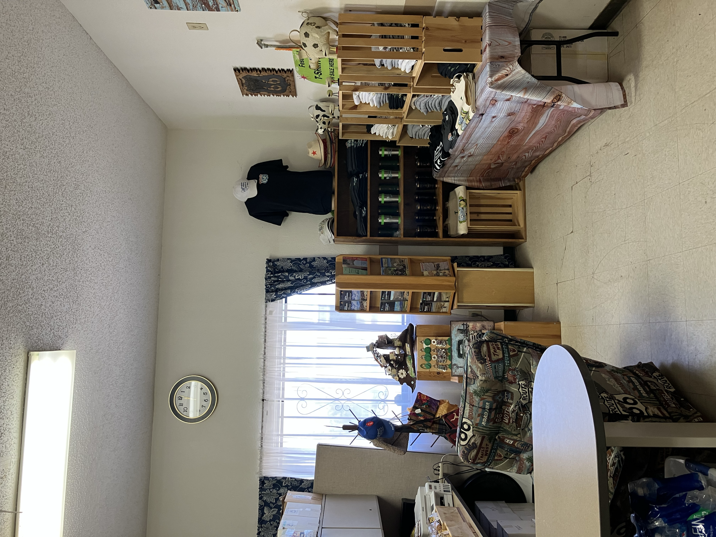 Room with a wall-mounted clock, a window with curtains, shelves with hats and sunglasses, a display of glass bottles, and tables covered with cloths displaying various items.