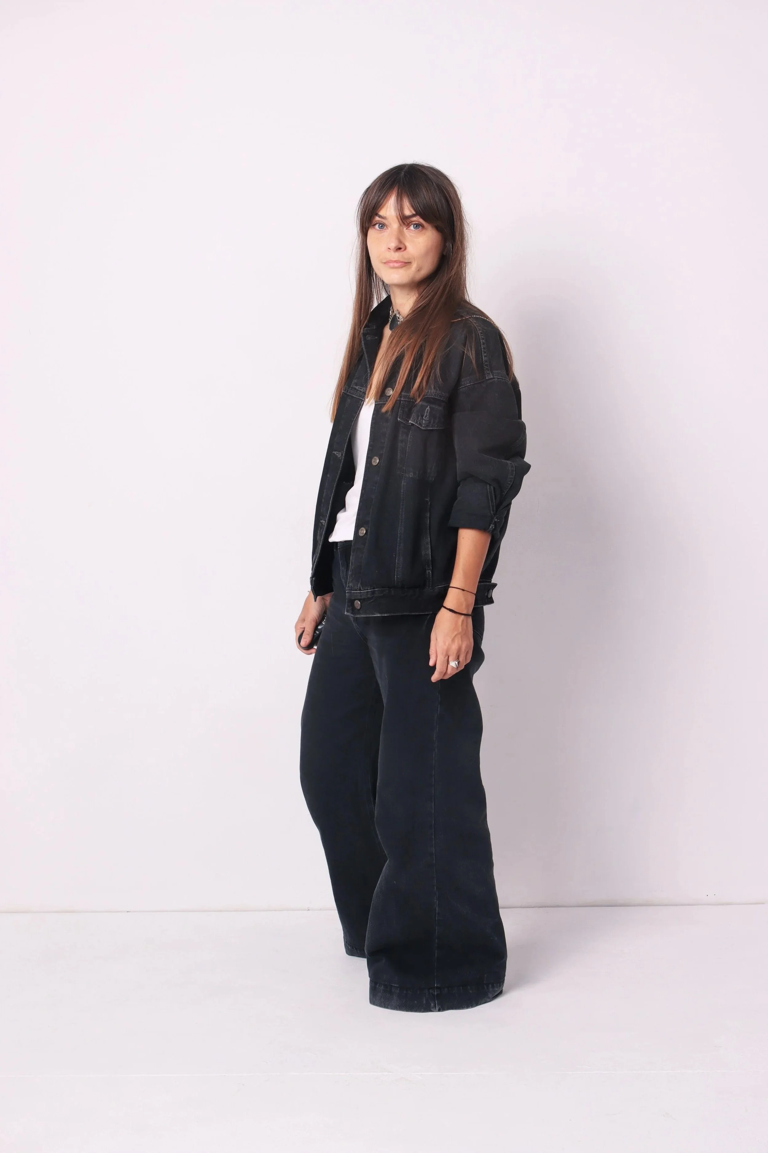 A woman with long brown hair and blue eyes stands against a plain white background, wearing a black denim jacket over a white shirt, with black wide-leg jeans, and holding a phone in her right hand.