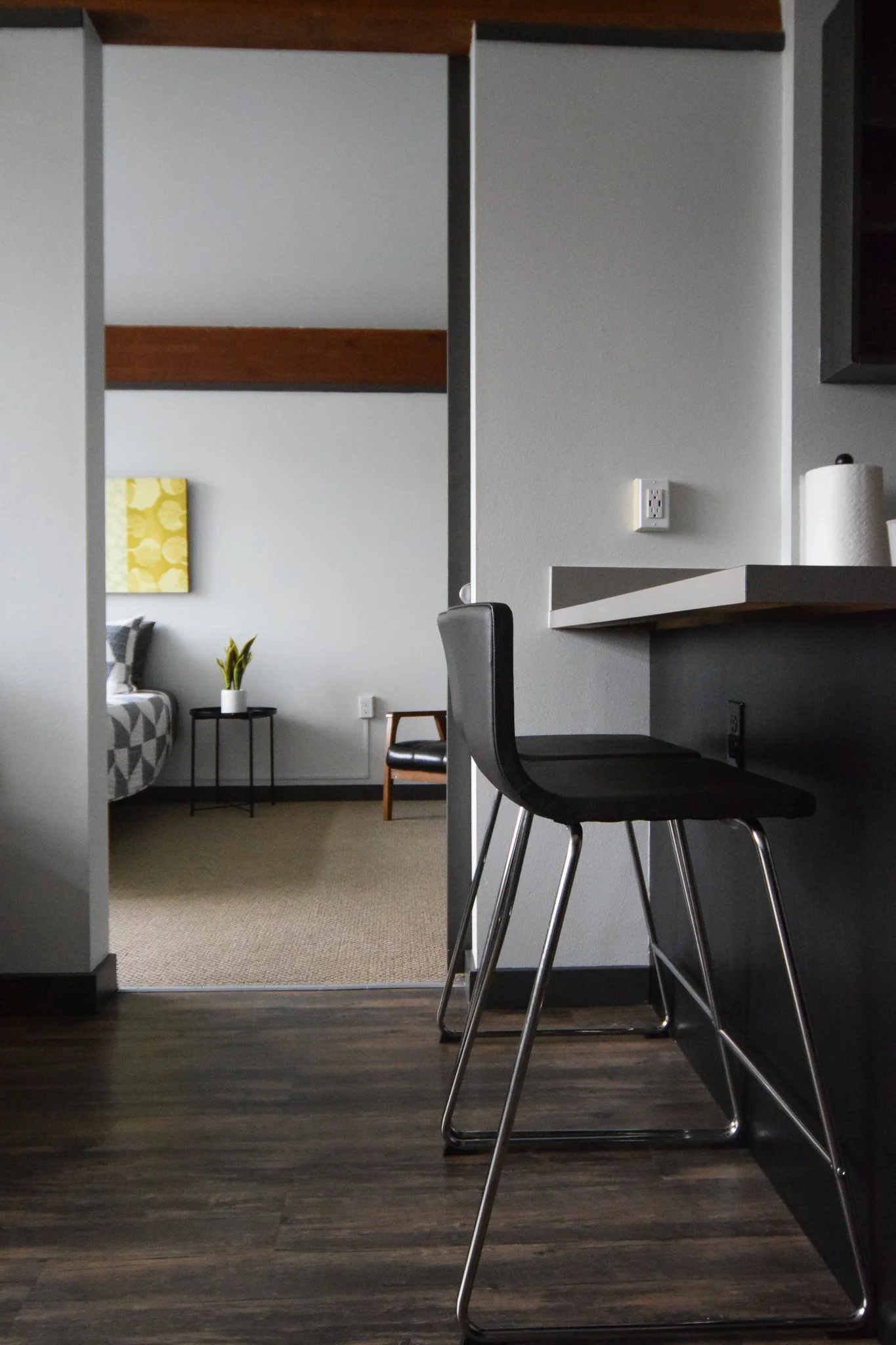 Interior view of a modern apartment with a dark wood floor, black bar stools at a white kitchen counter, and a living room in the background with a couch, a small table with a plant, and wall art.