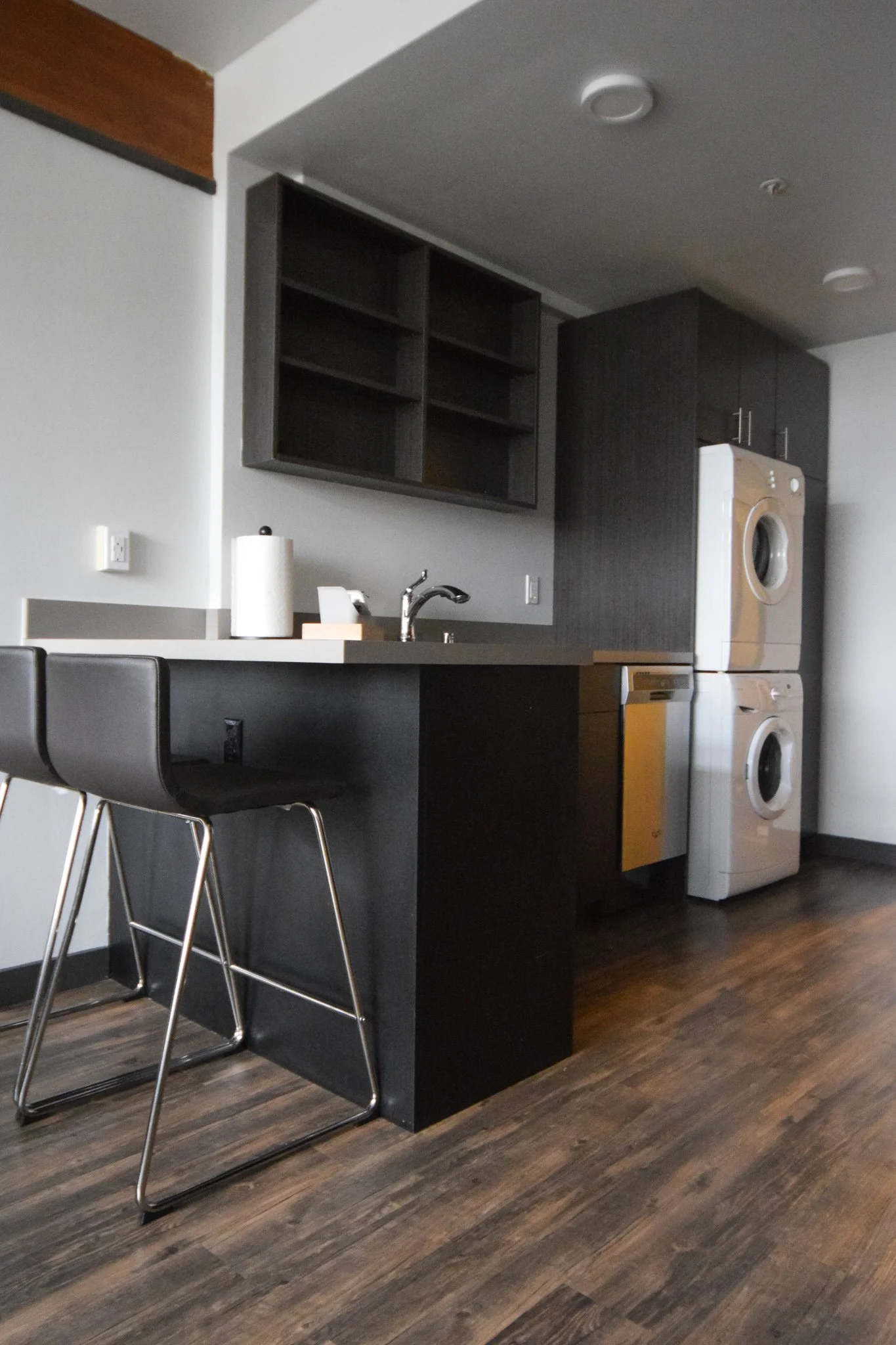 Modern kitchen with black cabinetry, stacked washer and dryer, and a bar-height counter with two black chairs.
