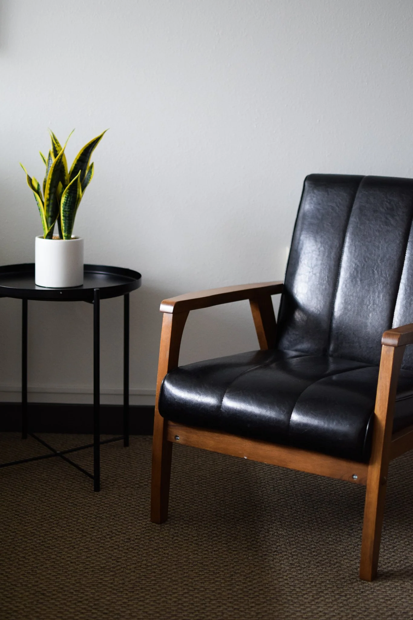 A black leather and wood armchair next to a small round black side table with a potted snake plant, on a brown textured carpet and plain white wall