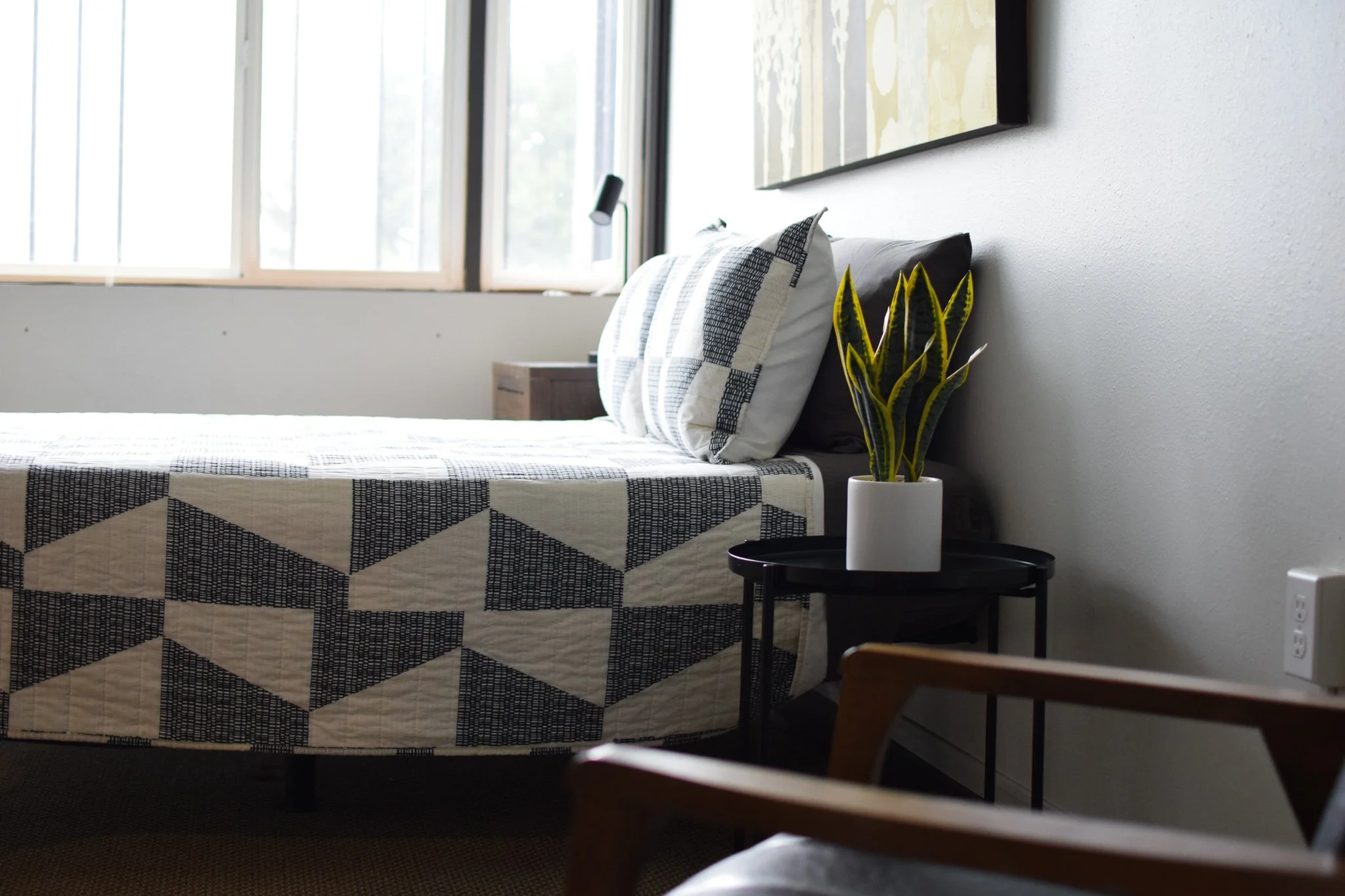 A bedroom with a bed featuring a geometric patterned quilt, two pillows, and a nightstand with a potted yellow-and-green snake plant, positioned near a window with natural light.