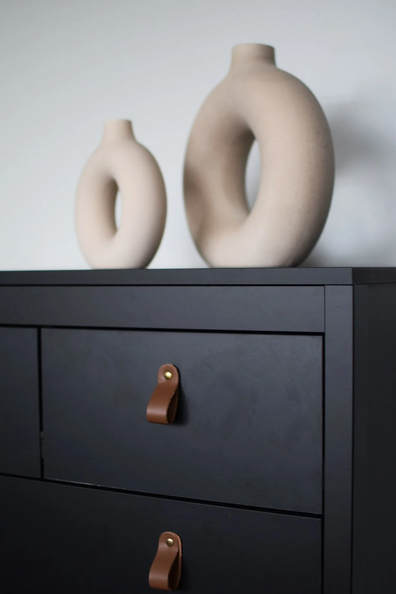 Two beige ring-shaped vases on top of a black dresser with brown leather handles.