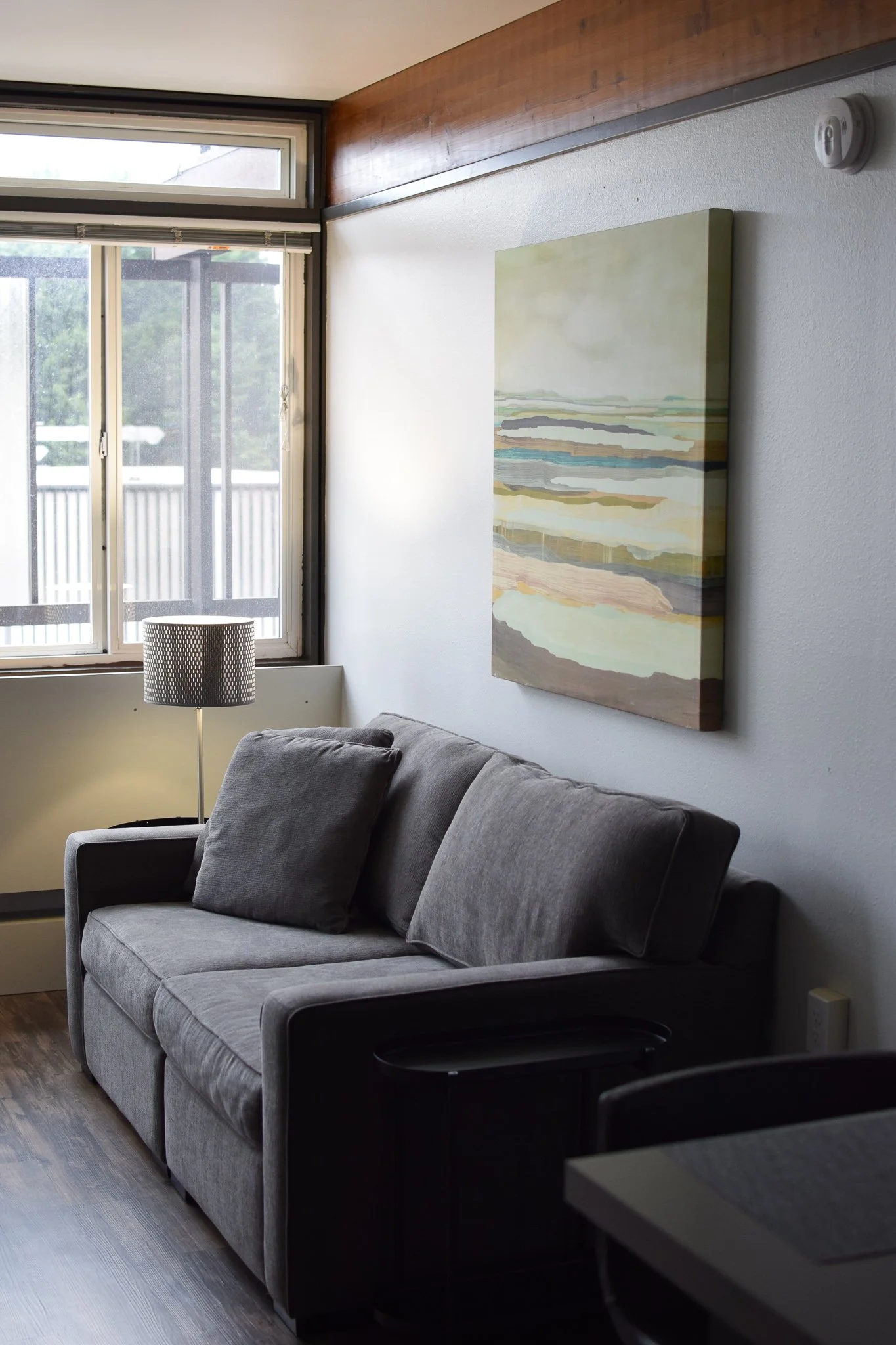 Living room with gray sofa, window with outdoor view, floor lamp, abstract landscape painting on wall.