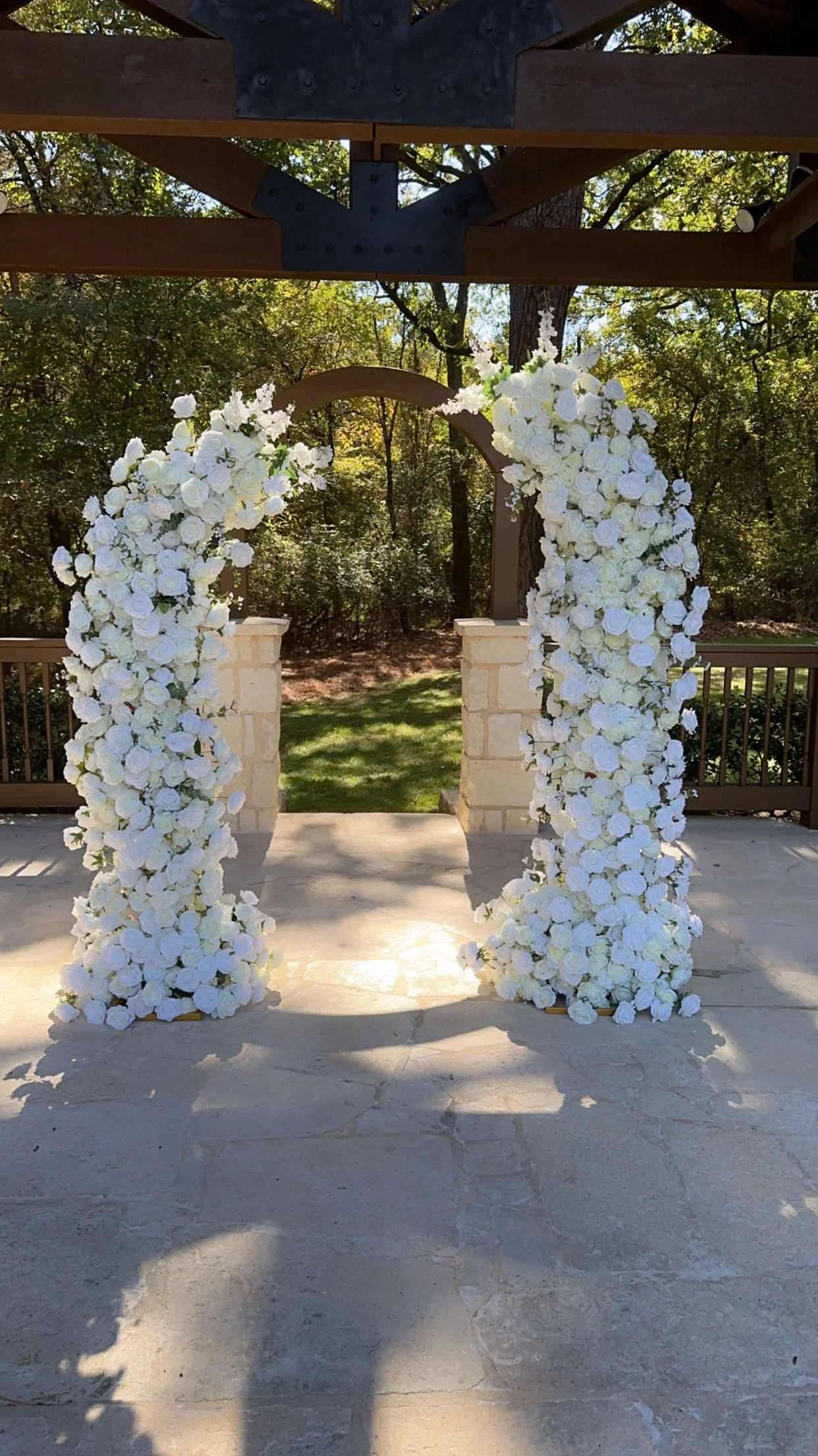 A wedding arch decorated with white roses and greenery, set outdoors against a backdrop of trees and a fence.