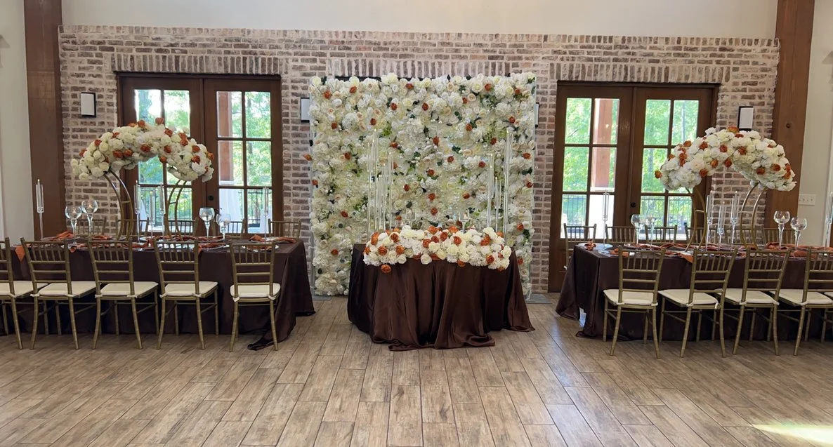 Wedding reception setup with long tables, floral centerpieces, and a floral backdrop behind a small table.