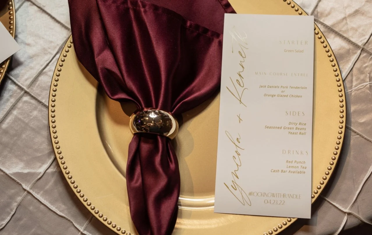 A table setting with a gold plate, a burgundy satin napkin tied with a gold ring, and a menu card with gold cursive writing.