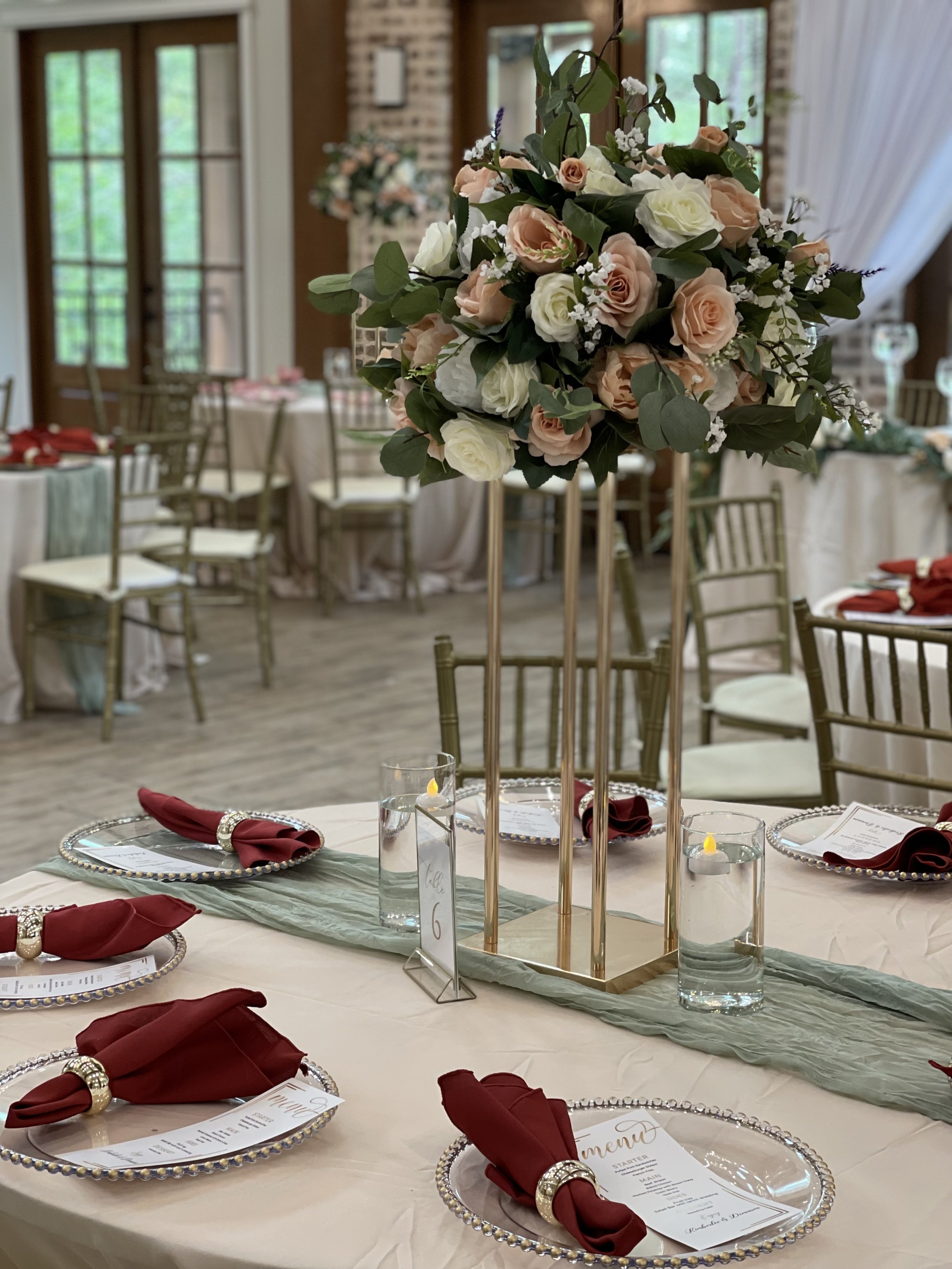 A decorated banquet table with a tall floral centerpiece featuring pink and white roses and greenery, set for a formal event with red napkins, menu cards, and glasses of water.