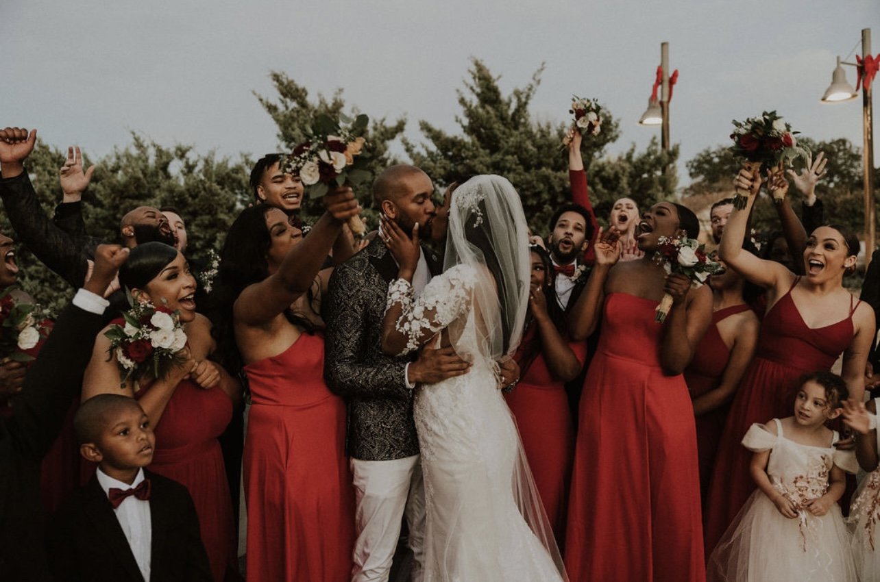 A wedding scene with the bride and groom kissing surrounded by bridesmaids and groomsmen celebrating, some holding bouquets and raising their hands in joy, outdoors with trees and decorative lighting in the background.