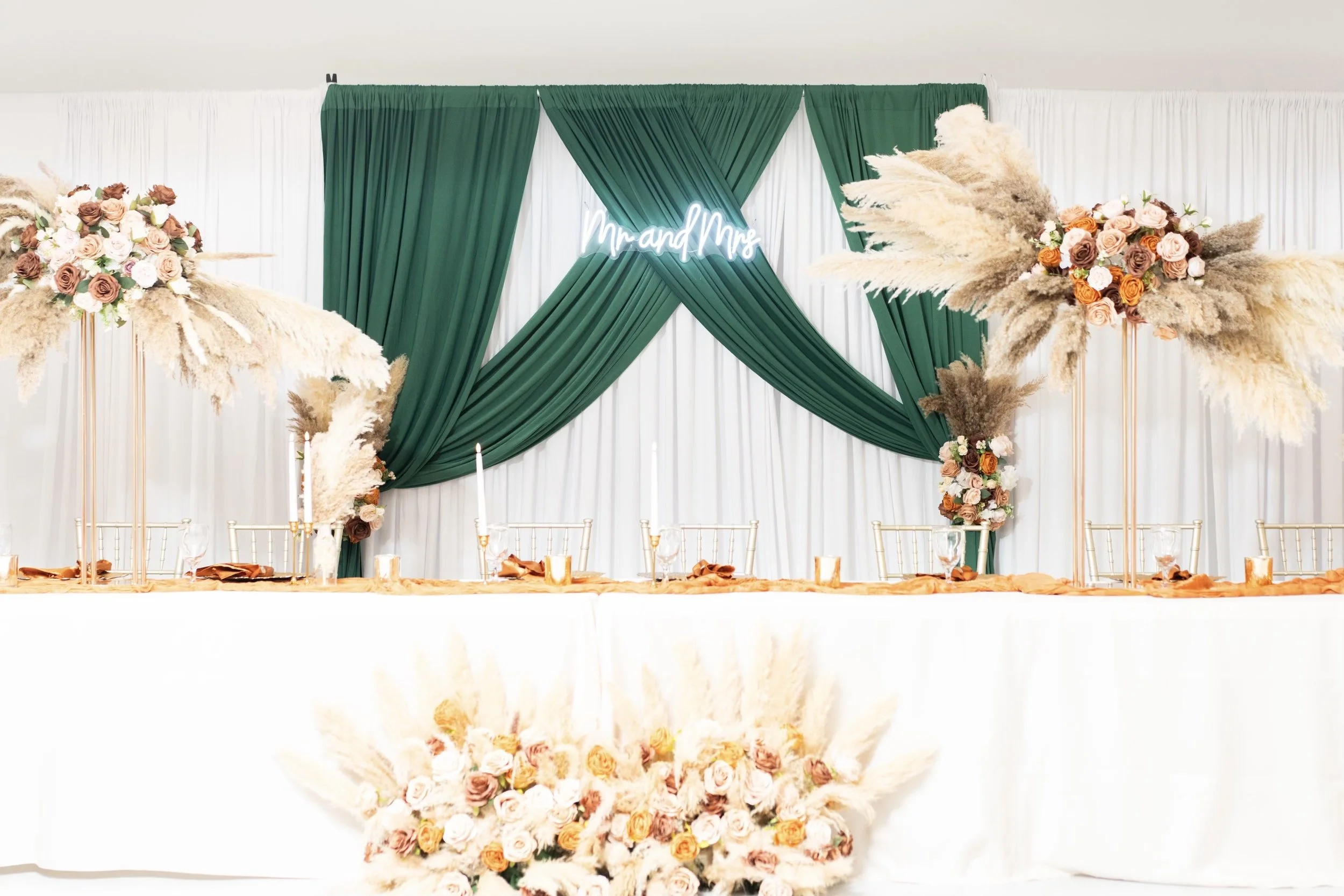 Wedding reception table decorated with floral arrangements and candles, with a backdrop of green and white drapes and a neon sign that says 'Mr and Mrs'.