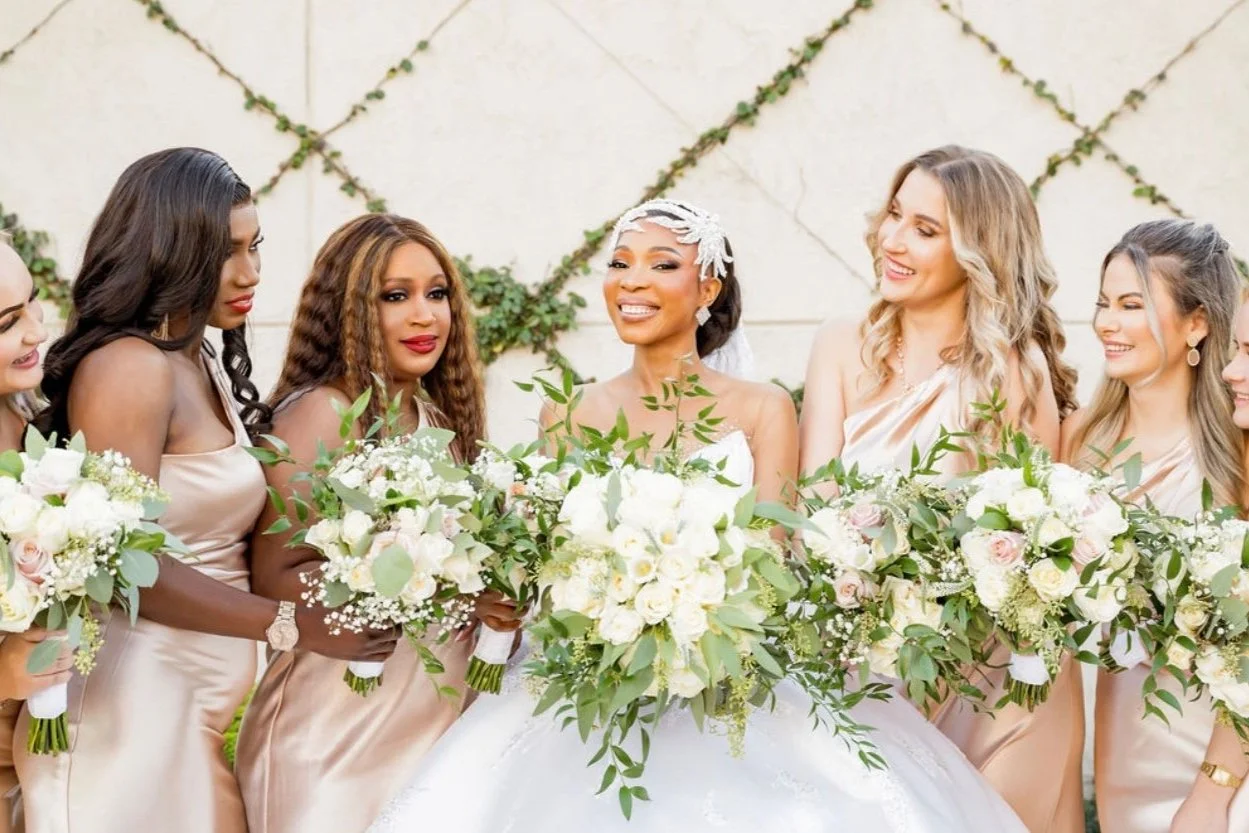 Group of women in a wedding celebration, holding bouquets of white flowers, standing outside against a wall with green vines, with the bride at the center in a white gown and floral headpiece.